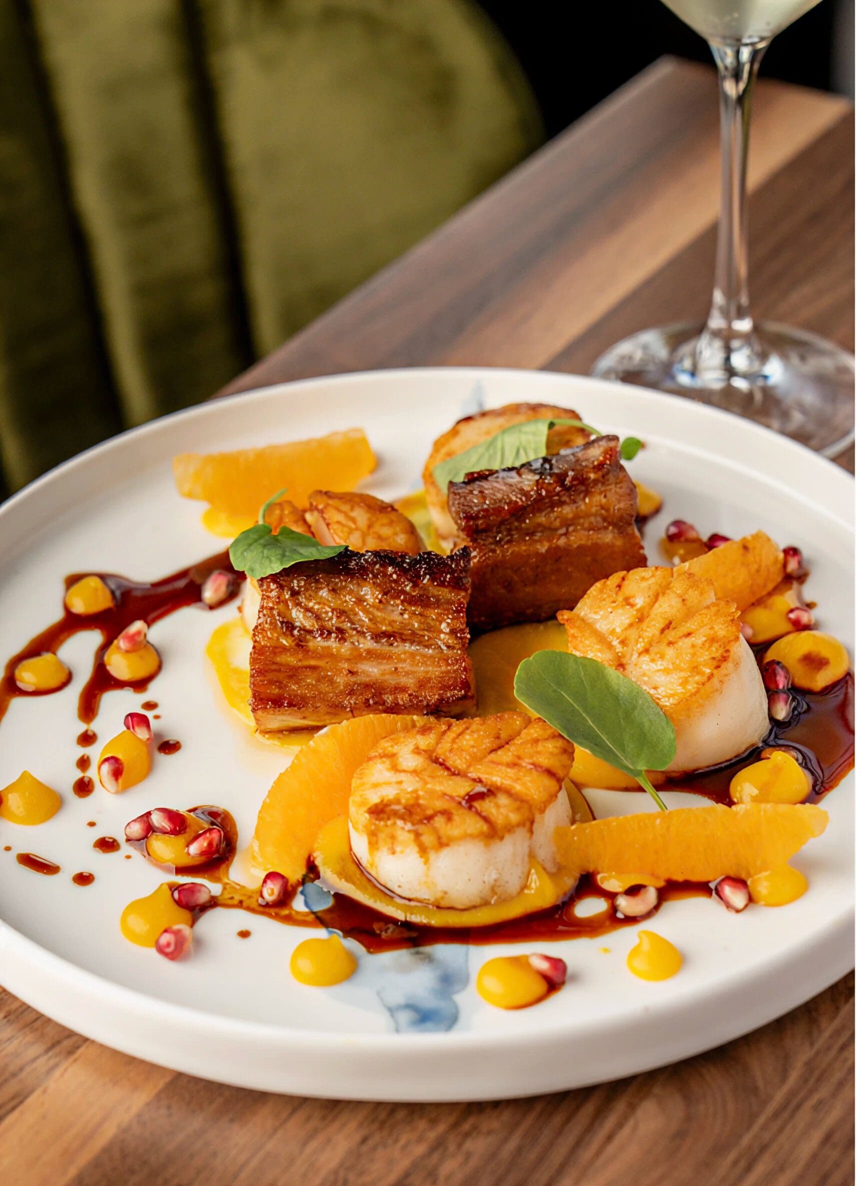 Seared scallops and pork belly on a white plate with orange segments, pomegranate seeds, and sauce drizzle.