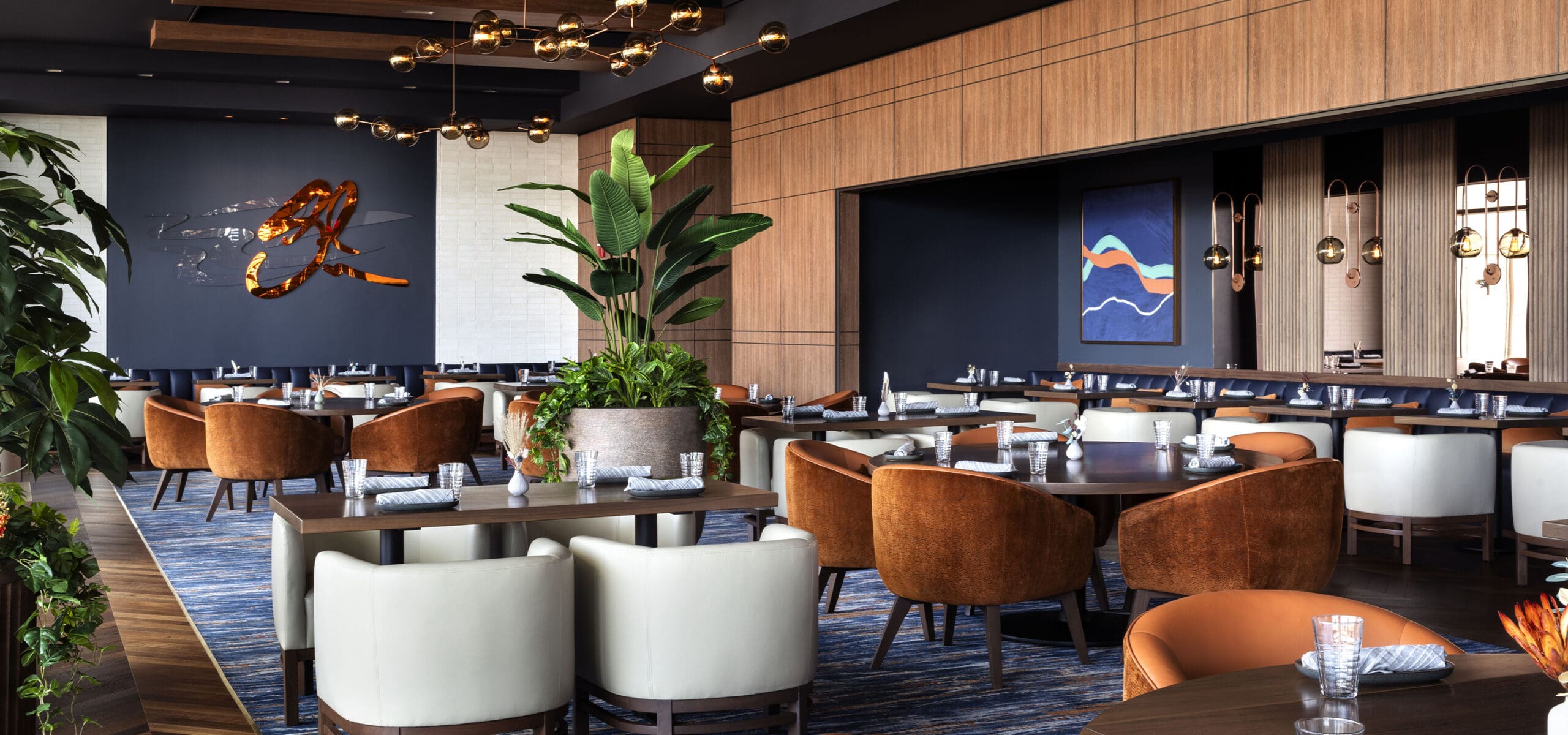 Modern restaurant with brown and white chairs, plants, wall art, and elegant lighting fittings.