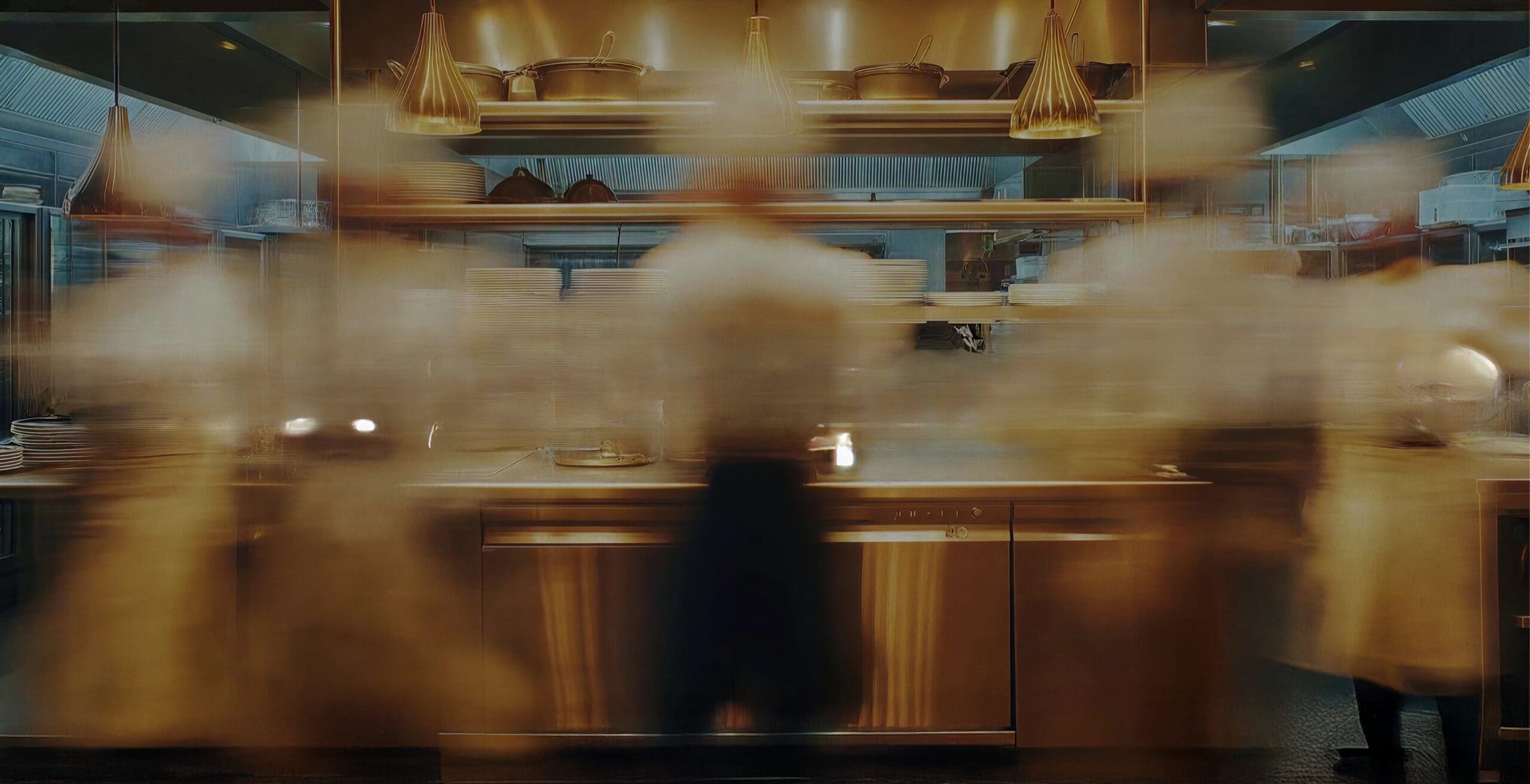 Blurred chefs move quickly in a modern, stainless-steel kitchen, creating a sense of speed and activity.