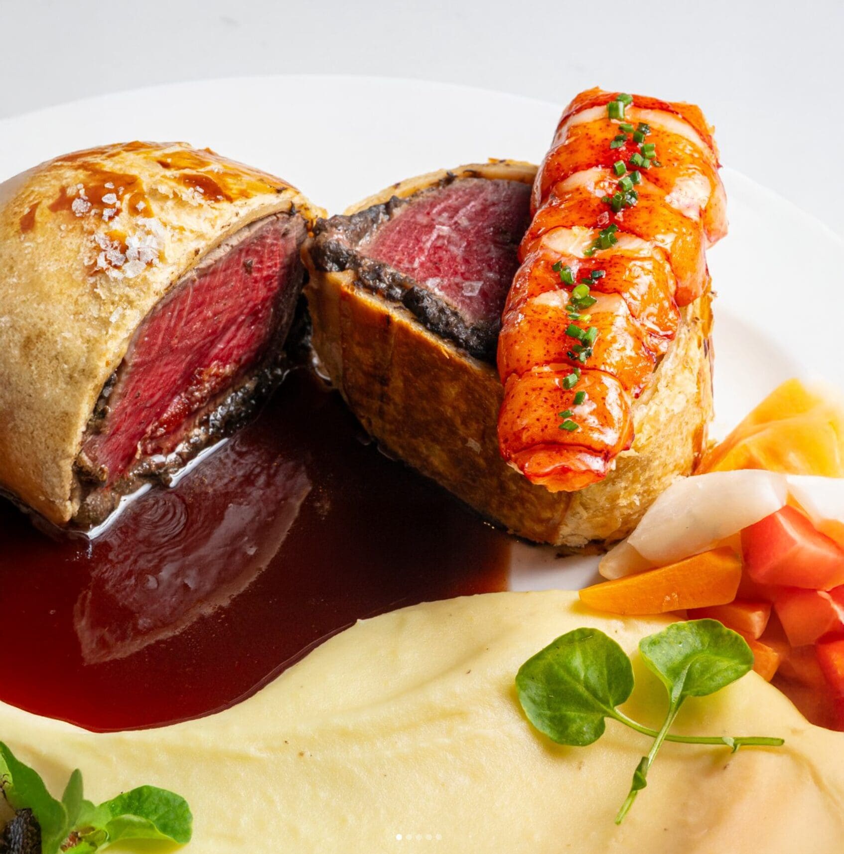 Beef Wellington with lobster, mashed potatoes, root vegetables, greens, and a rich sauce on a white plate.