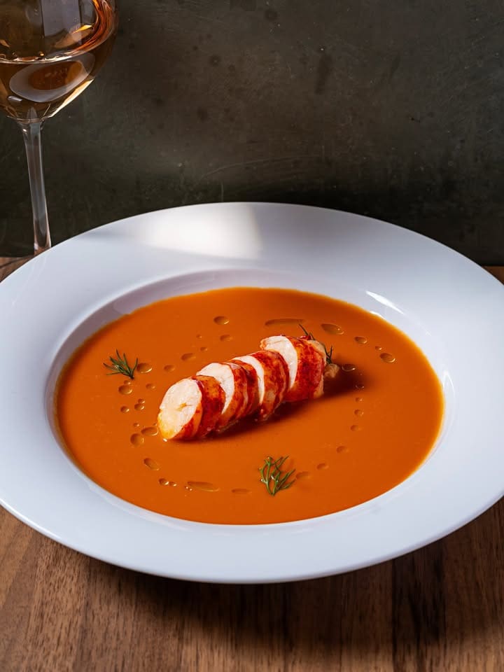 White plate of orange soup with sliced lobster tail, garnished with herbs, next to a glass of white wine.