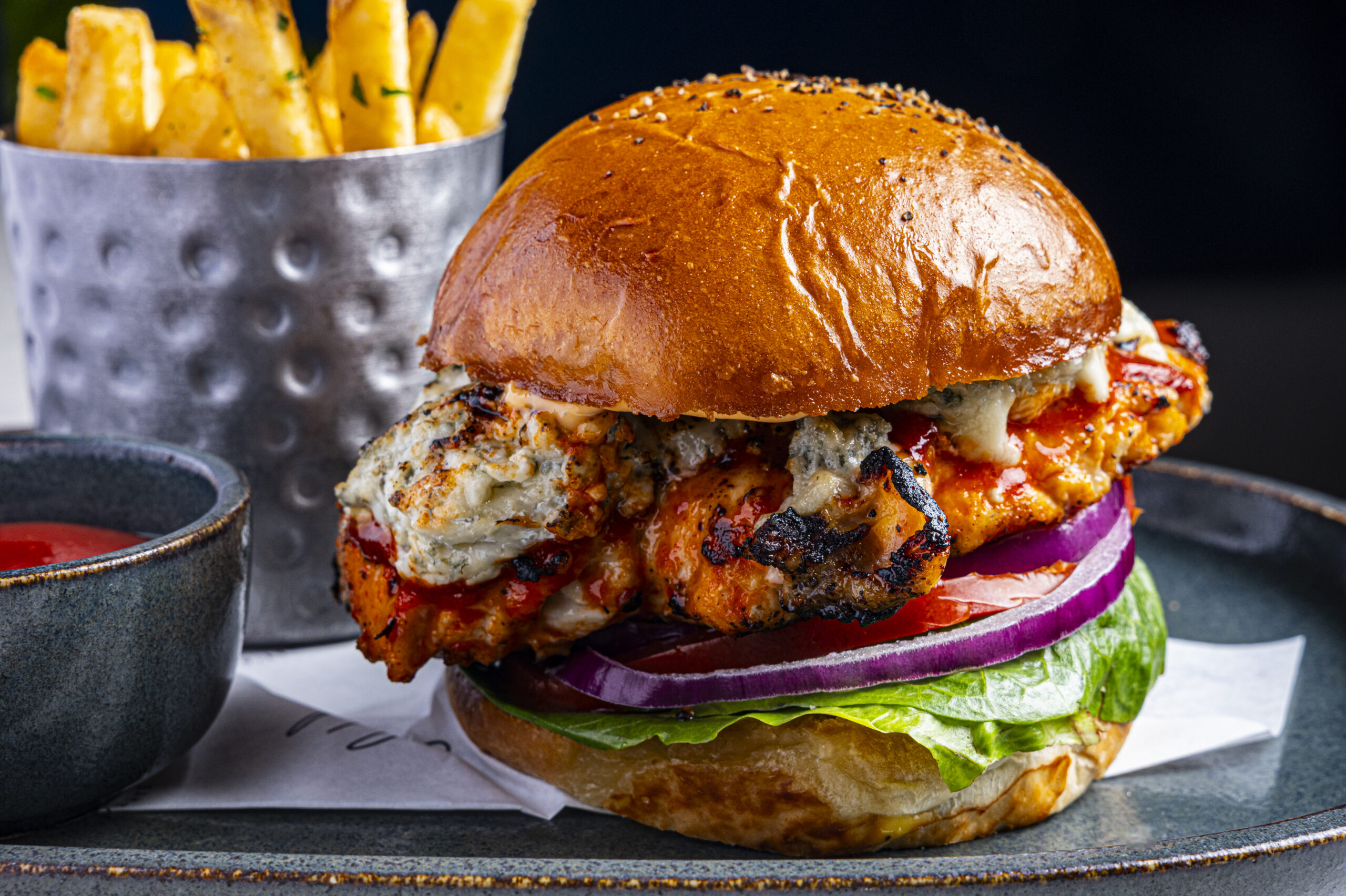 A grilled chicken sandwich with blue cheese and sauce, served with a side of chips and dipping sauce.