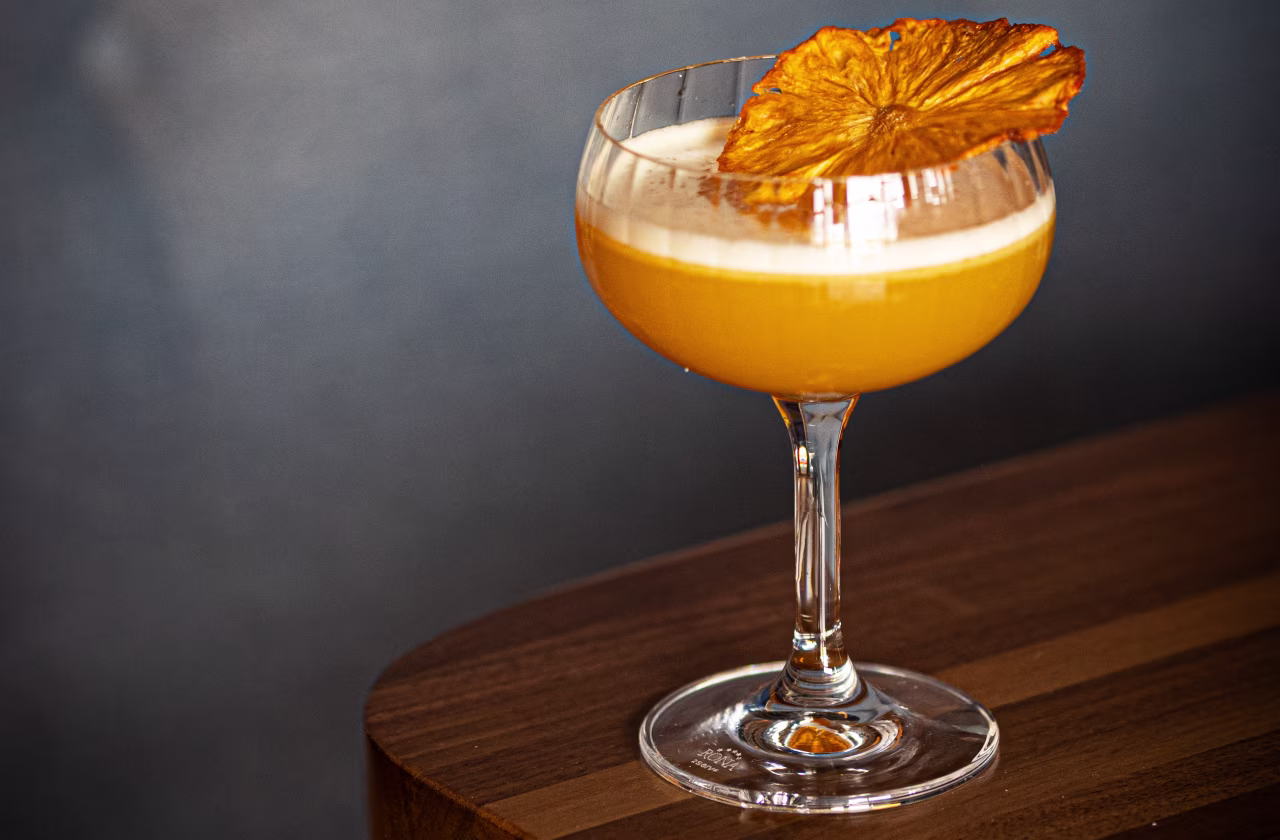 A yellow cocktail in a coupe glass with froth and a dried pineapple garnish on top, on a wooden table.