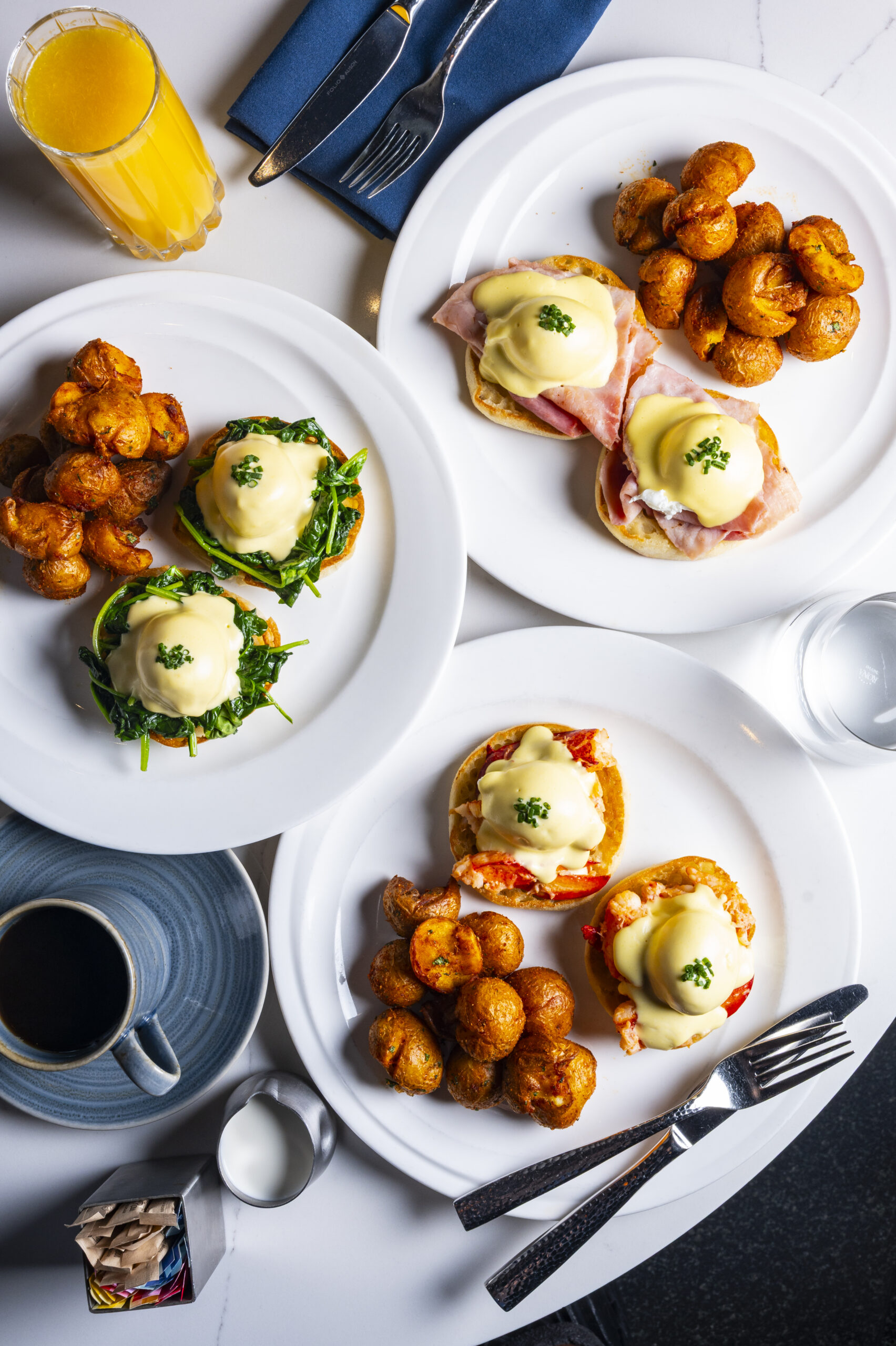 Three plates of eggs Benedict served with roast potatoes and a glass of orange juice.