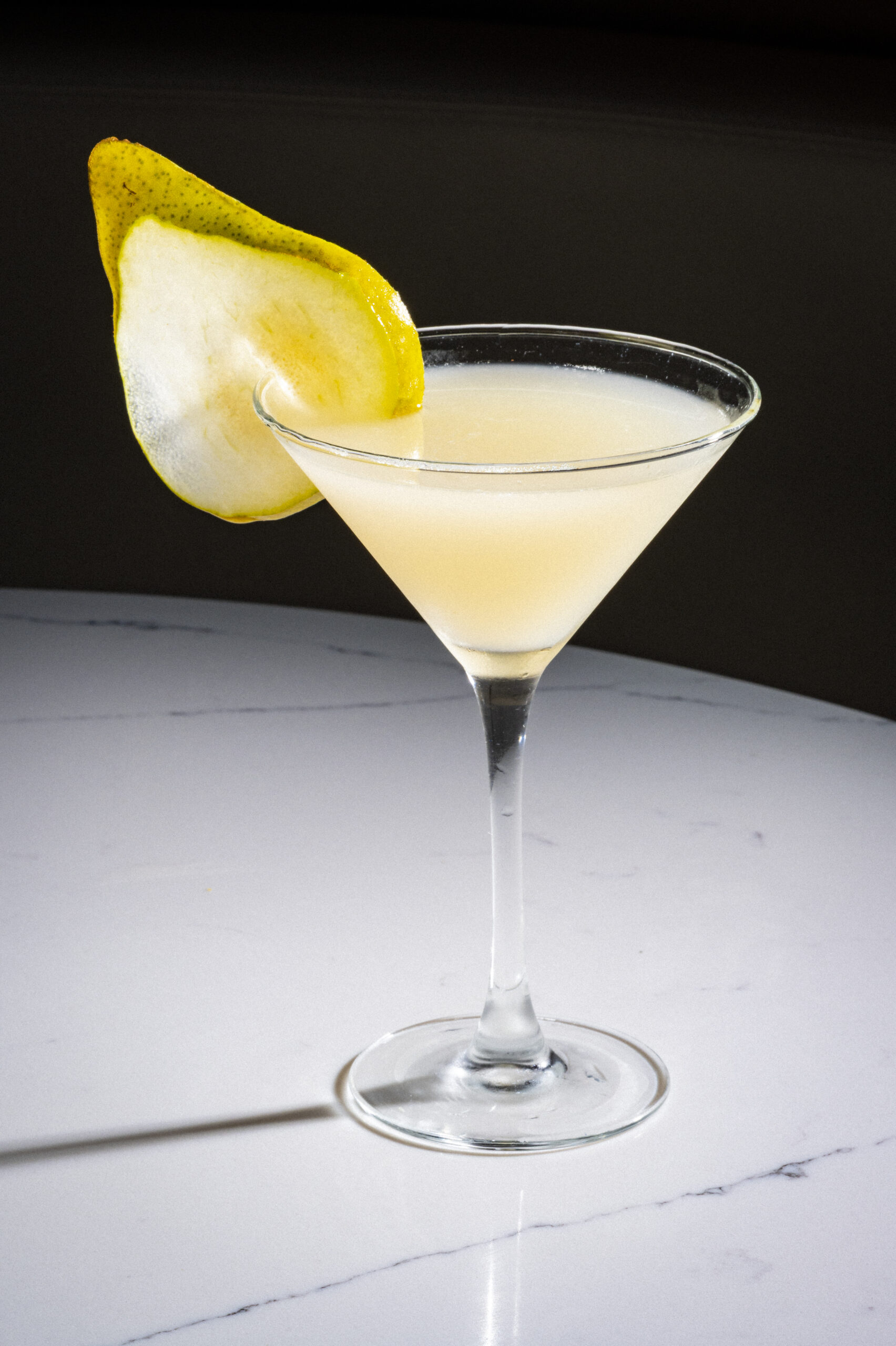 A light-coloured cocktail in a martini glass garnished with a pear slice.