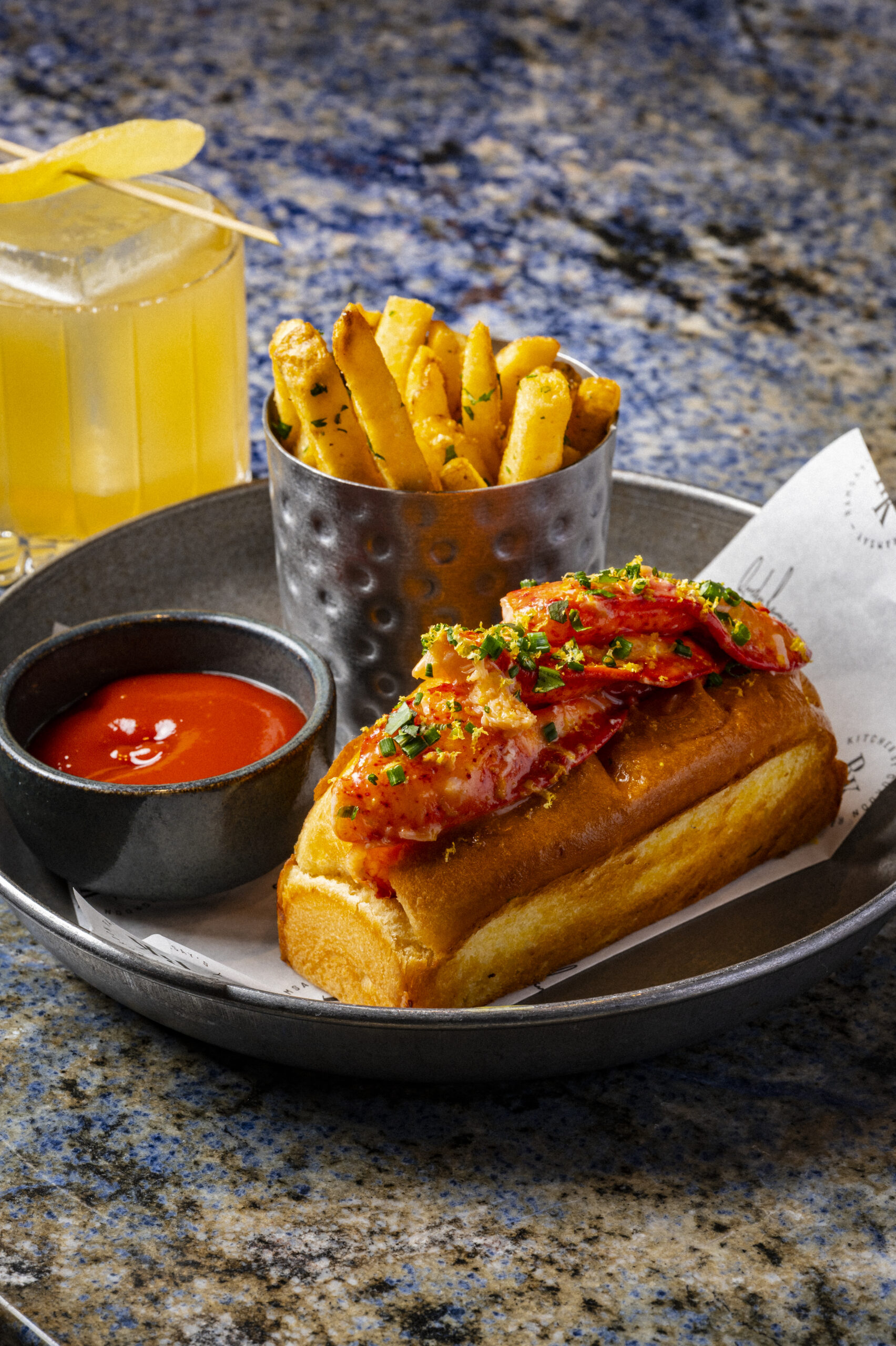 Lobster roll with a side of chips, tomato sauce, and a yellow cocktail.