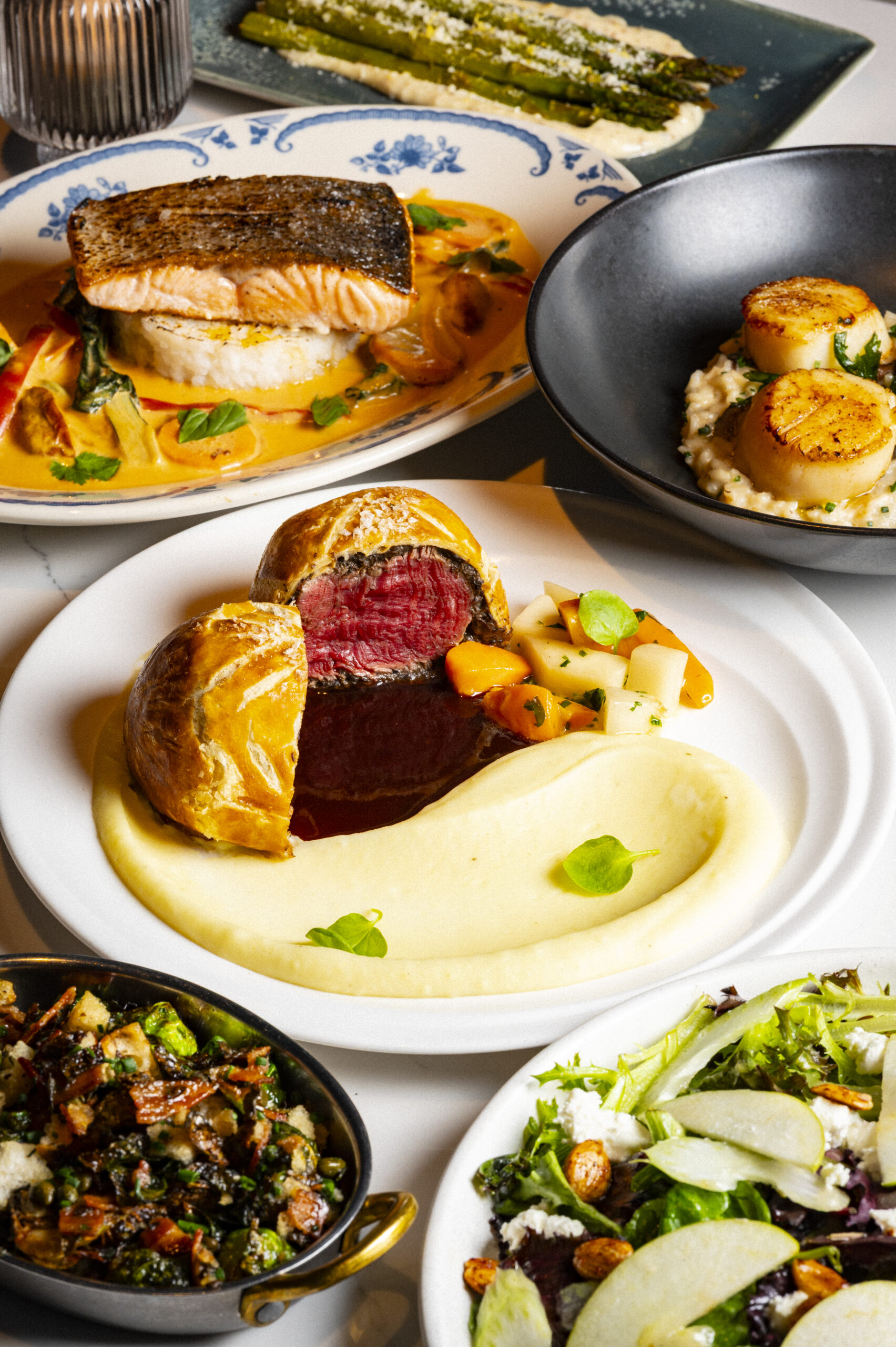 A spread of dishes including beef Wellington with mashed potatoes, seared scallops, salmon, asparagus, and salad.