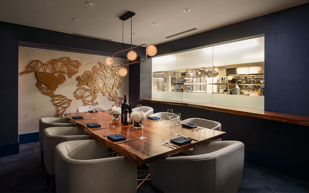 A live-edge wooden dining table set for dinner in front of a window looking into a kitchen.