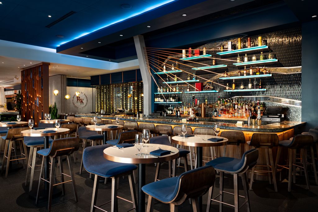 Restaurant interior featuring round tables with blue chairs facing a backlit bar with floating shelves.