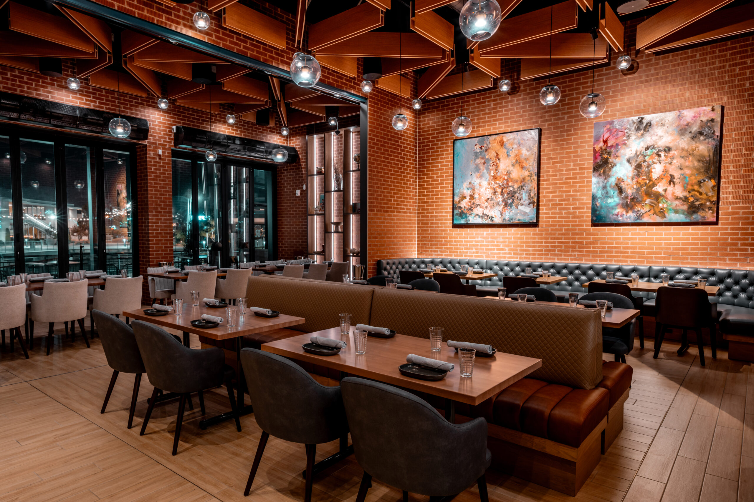 Restaurant interior with wooden tables, gray upholstered chairs, and banquettes against a brick wall with abstract art.