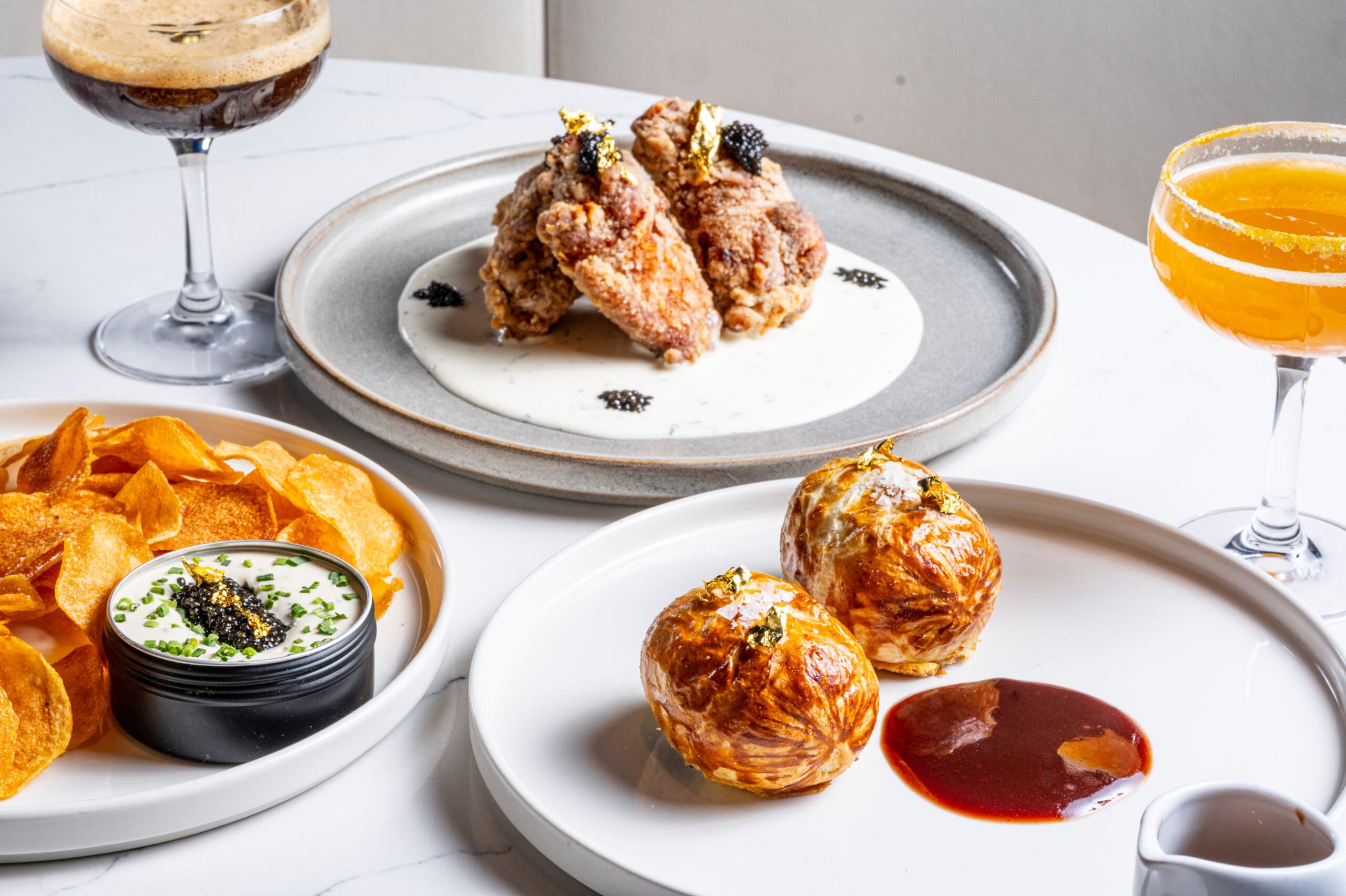 Fried chicken on sauce, fried potato balls, crisps with dip, and cocktails on a white table.