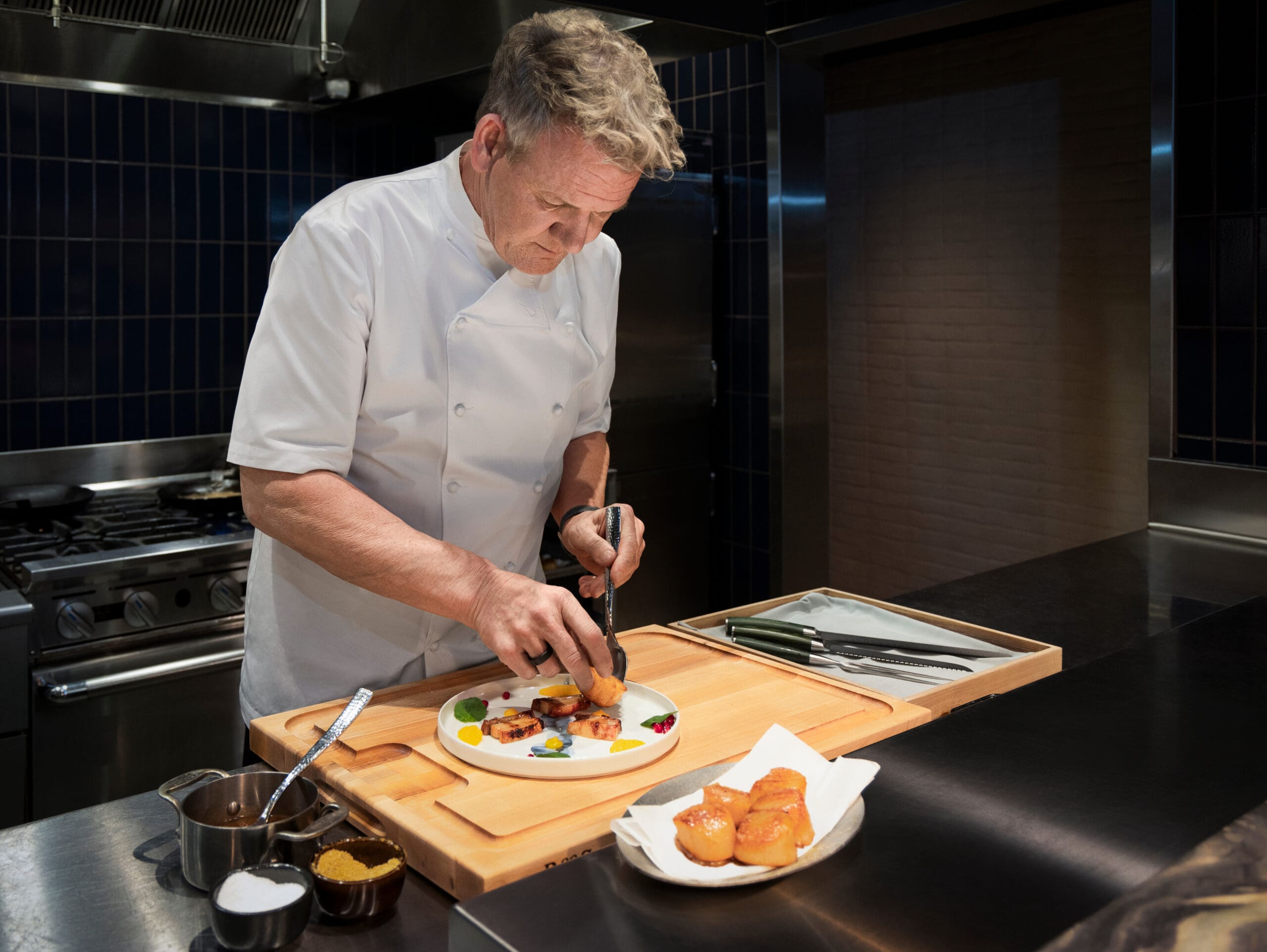 Gordon Ramsay in a white coat carefully plating food in a modern kitchen with utensils nearby.