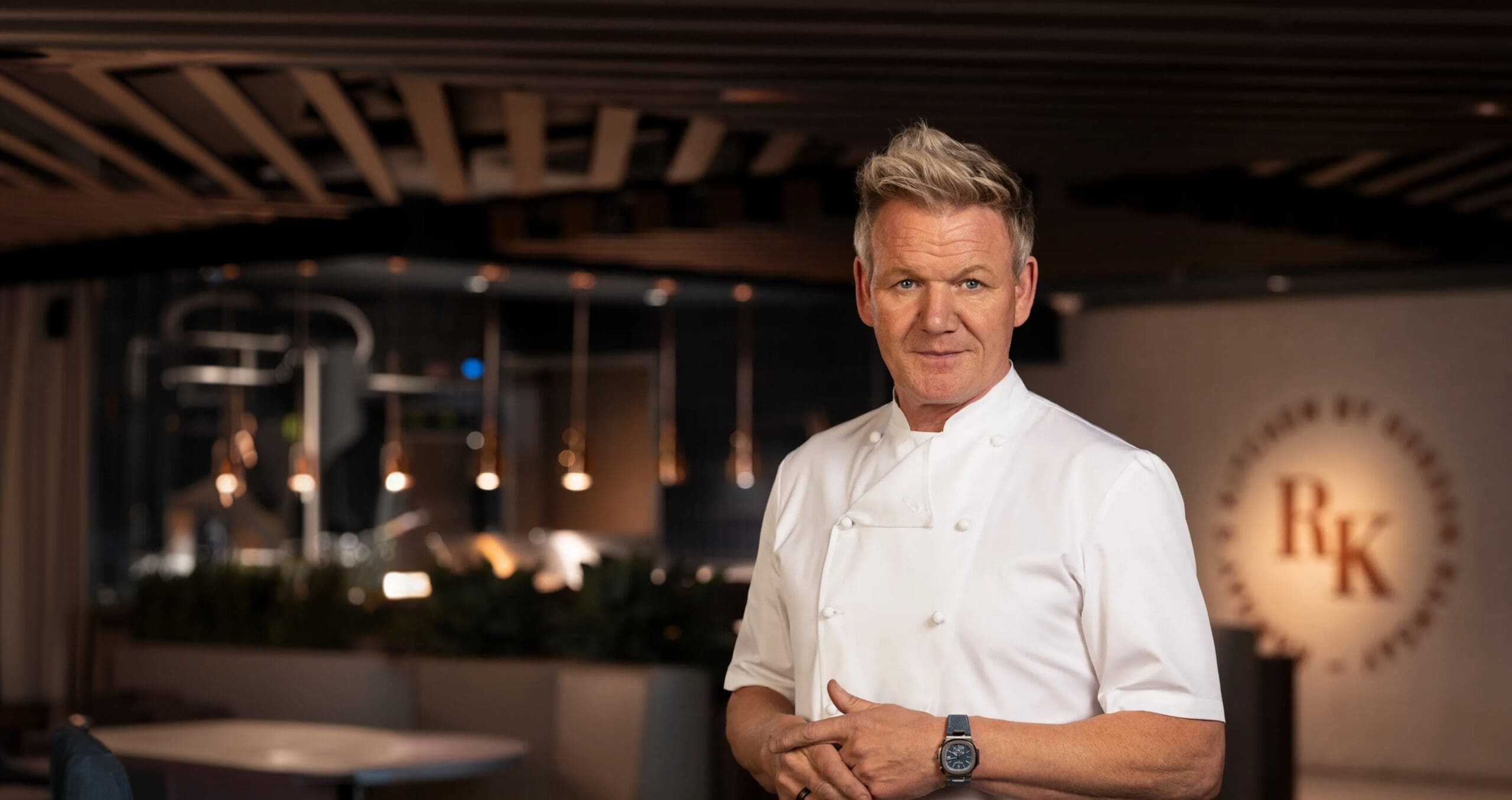 Gordon Ramsay in a white uniform stands confidently in a modern restaurant with pendant lights in the background.