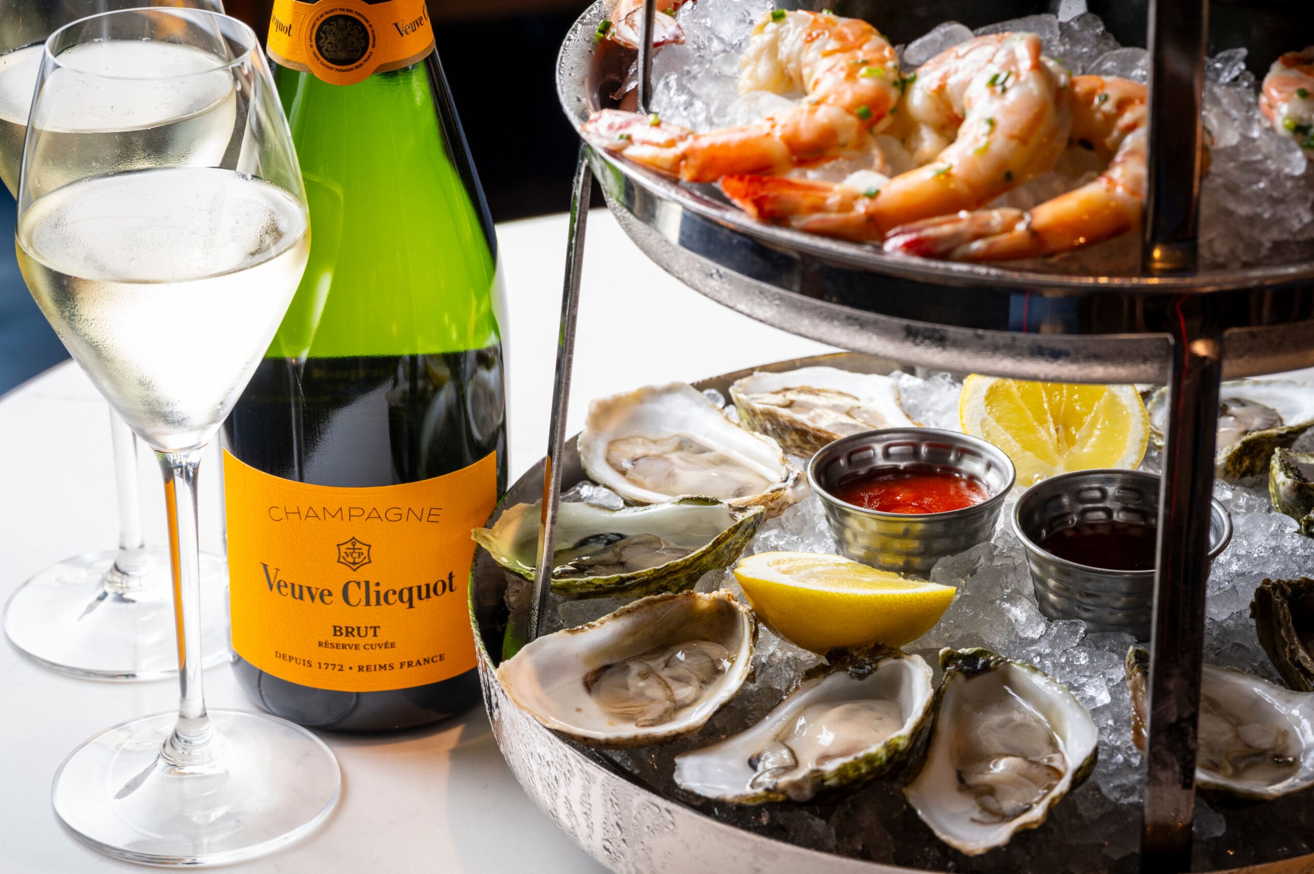 Champagne, oysters, prawn cocktail, and lemon wedges on ice, with two glasses of fizz on a white table.