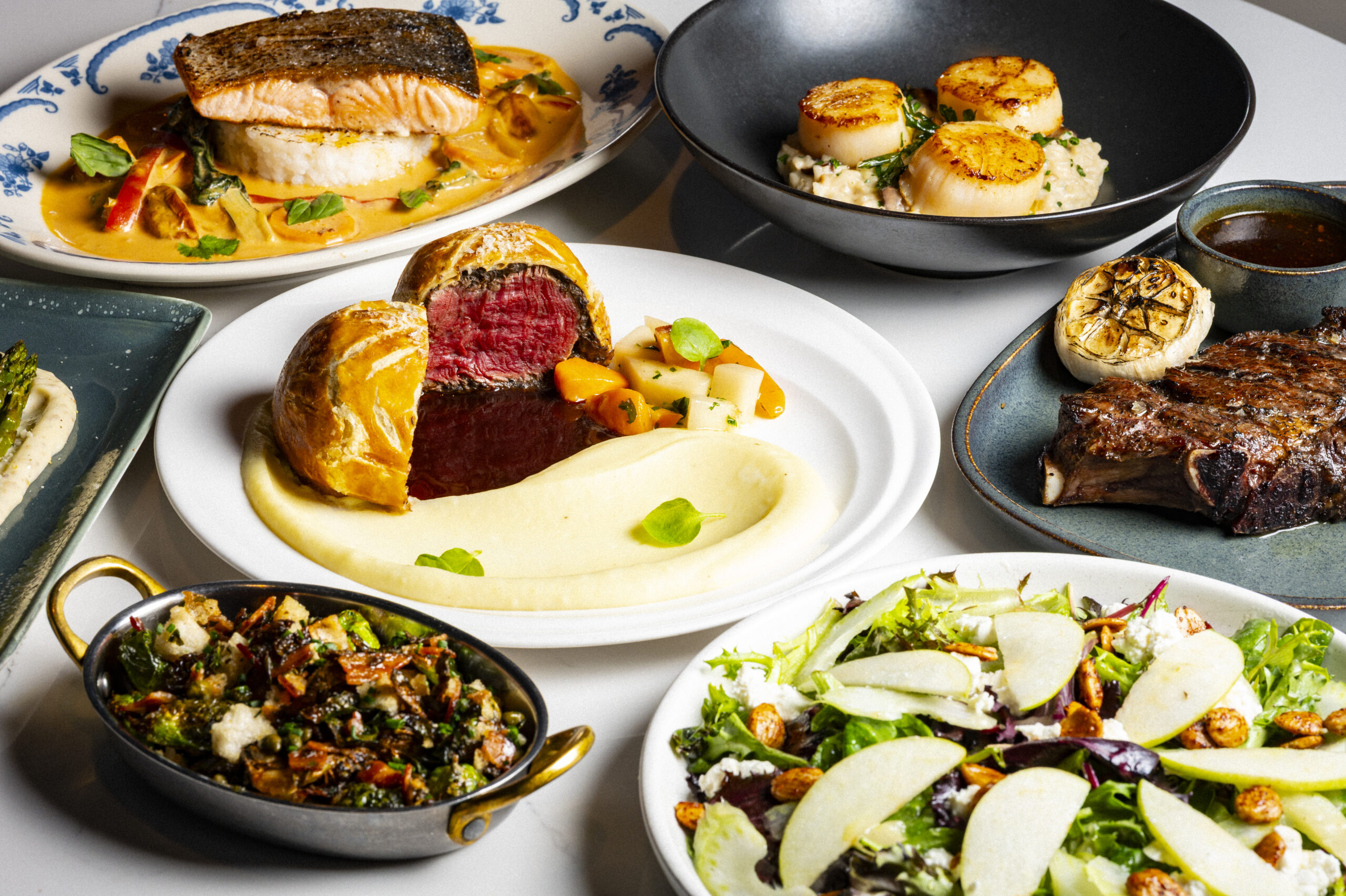 A table spread featuring salmon, seared scallops, beef Wellington, steak, roasted vegetables, and a salad.