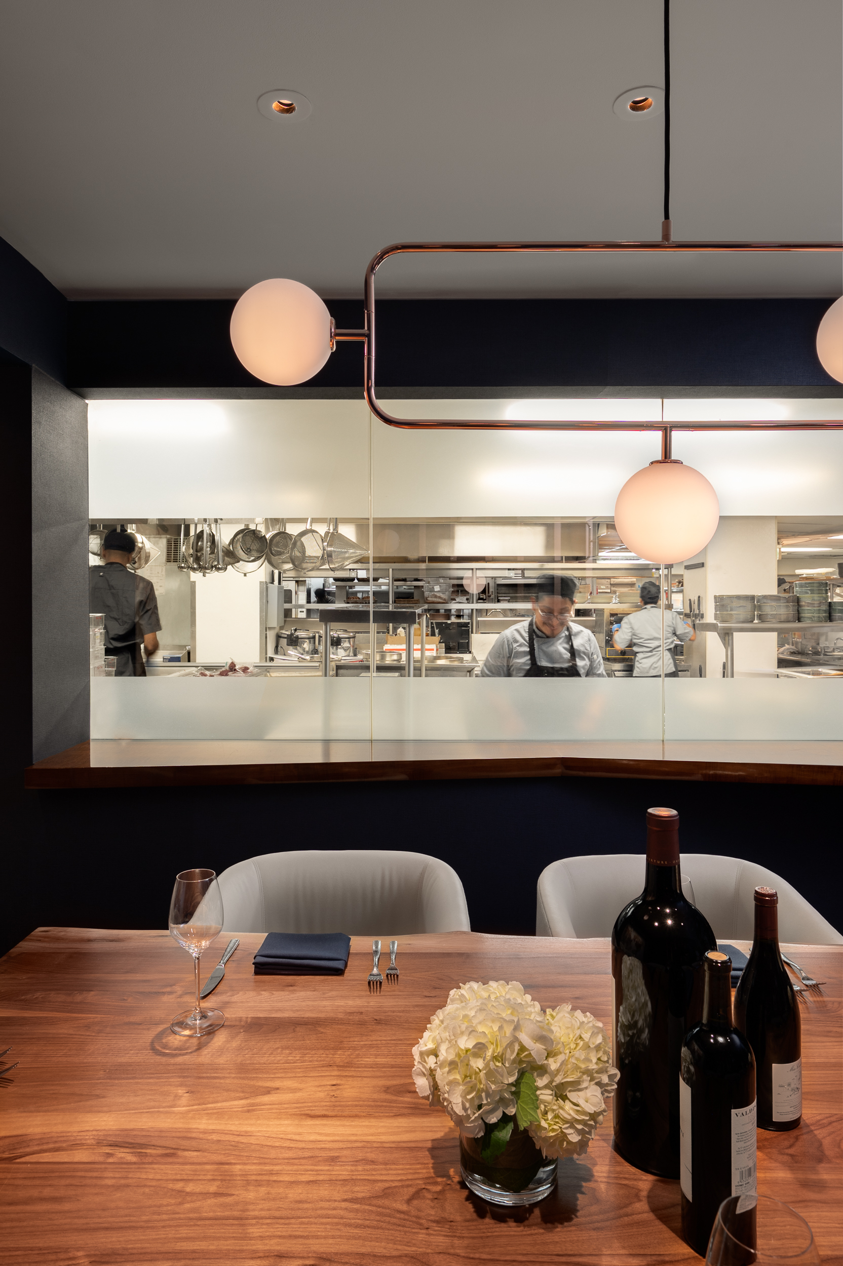 Dining table with wine bottles and flowers in front of an open restaurant kitchen.