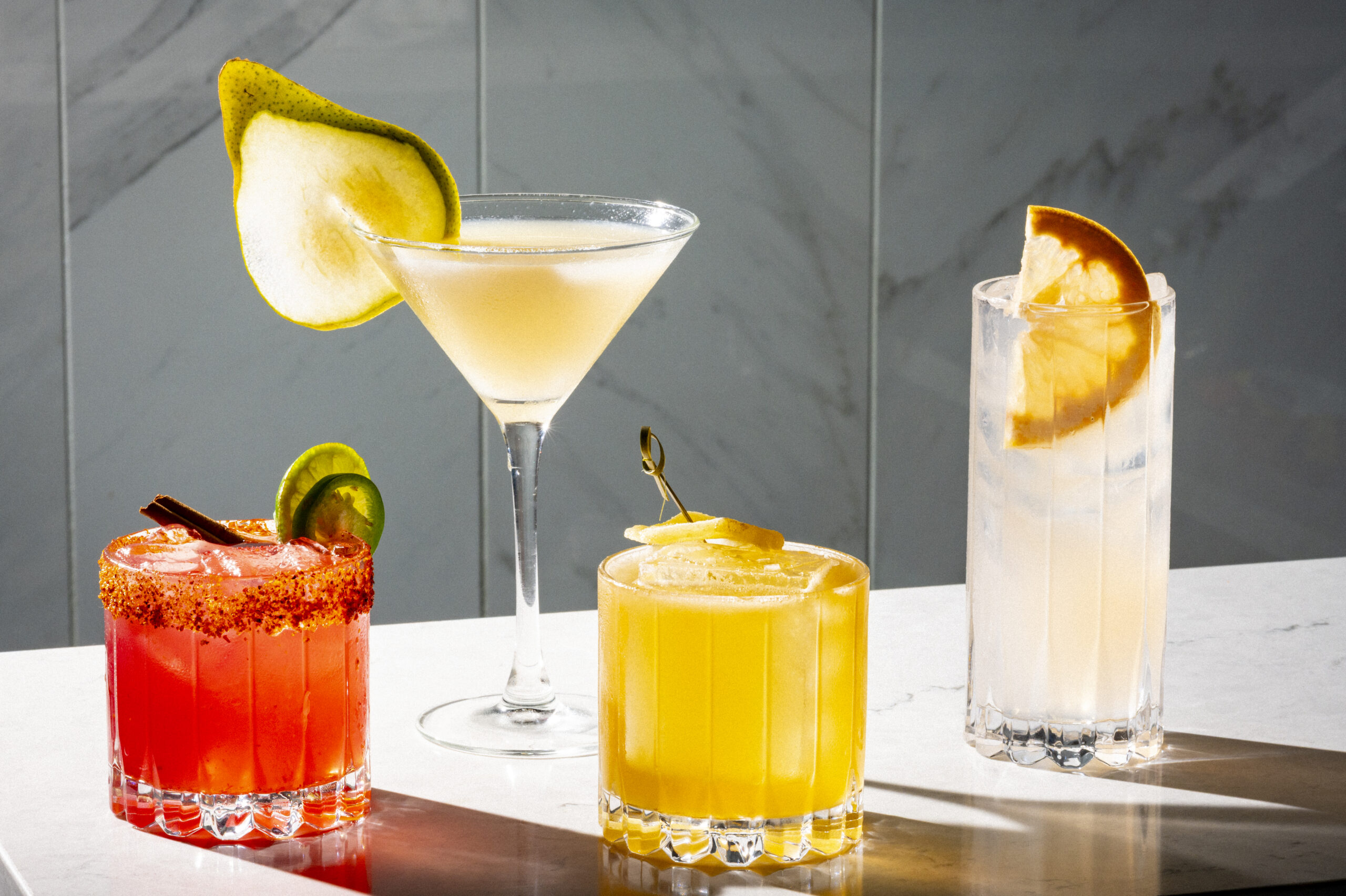Four cocktails on a table, including a spicy margarita, a pear martini, a yellow drink, and a citrus highball.
