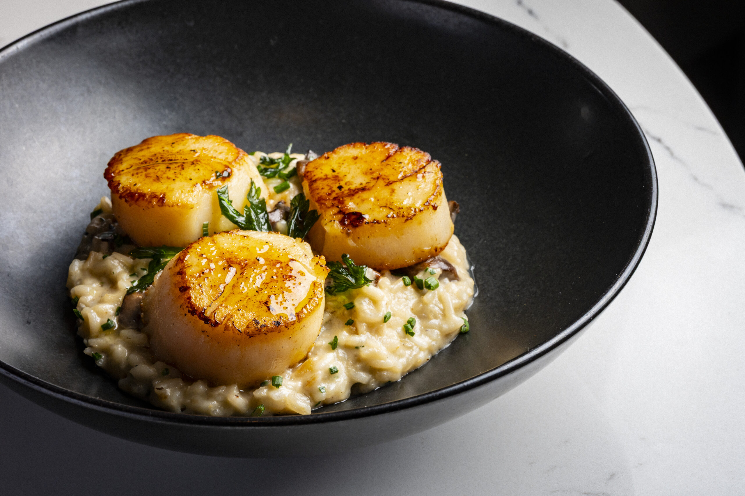 Three seared scallops served over a bed of creamy risotto in a black bowl.