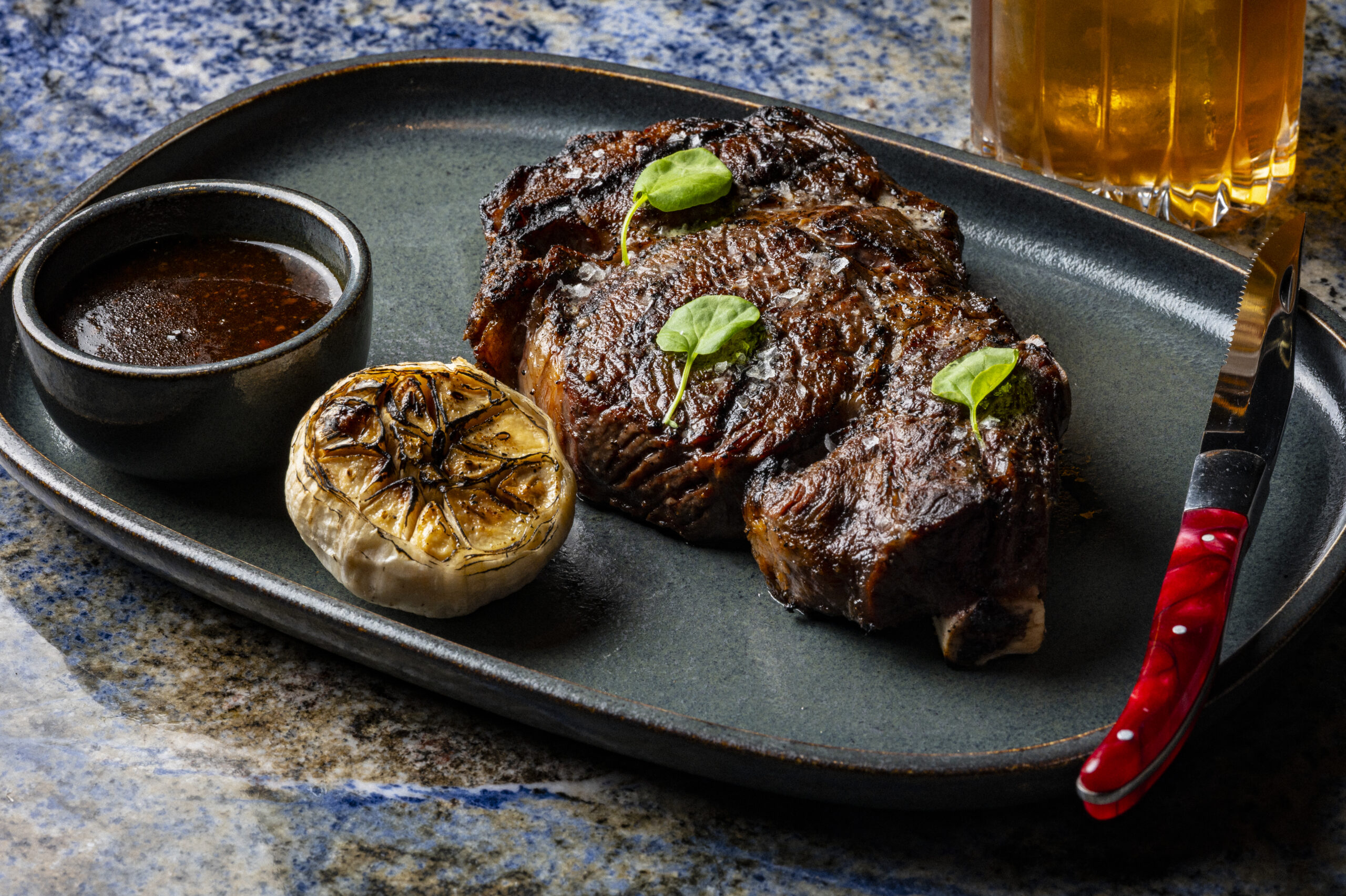 Grilled steak garnished with herbs, served with roasted garlic and a side of sauce on a dark platter.