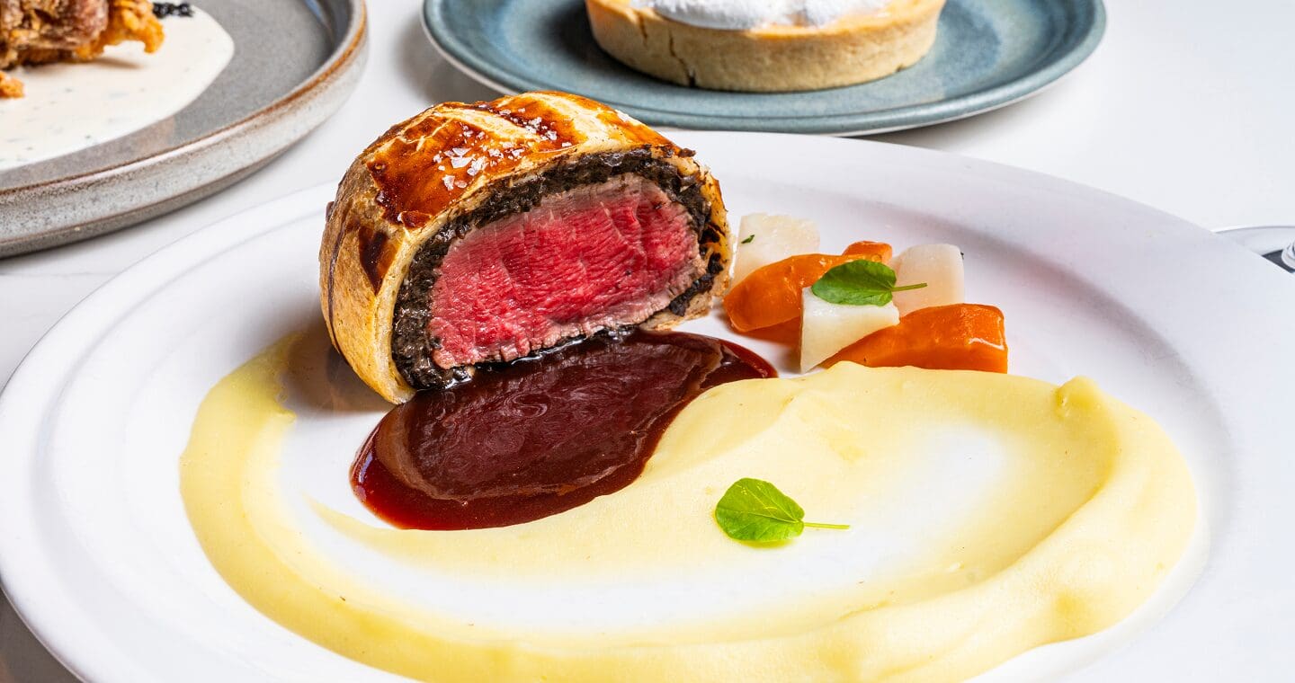 Sliced beef Wellington with sauce, creamy mashed potato, and vegetables on a white plate.