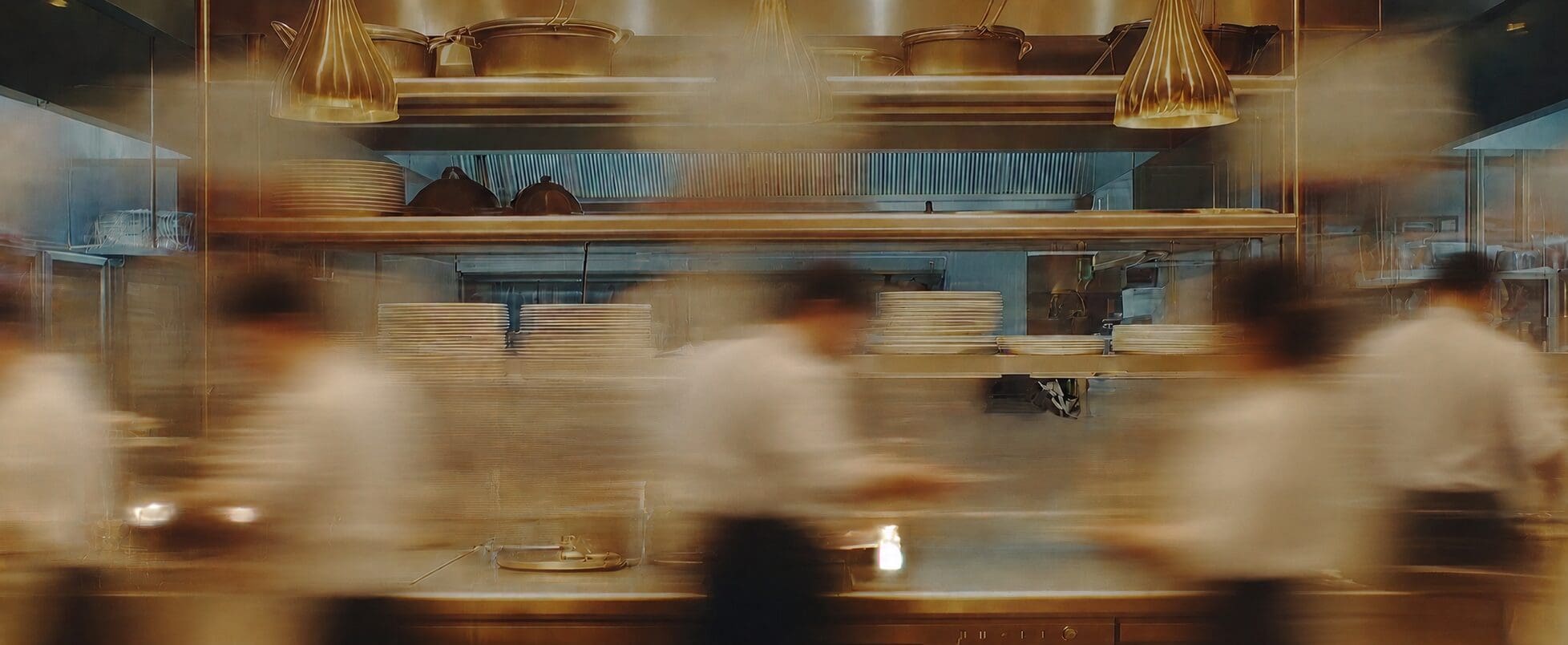Busy restaurant kitchen with blurred staff moving quickly, metal shelves, and stacks of white plates.