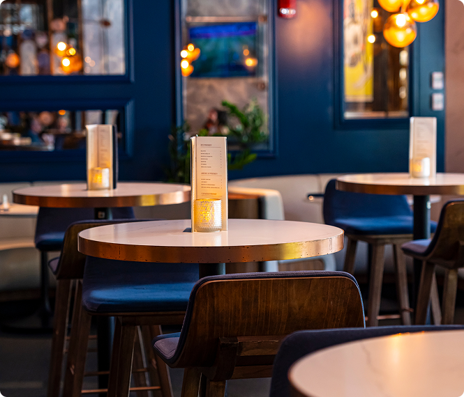 Round tables with menus and candles, surrounded by wooden chairs in a cozy, modern restaurant.