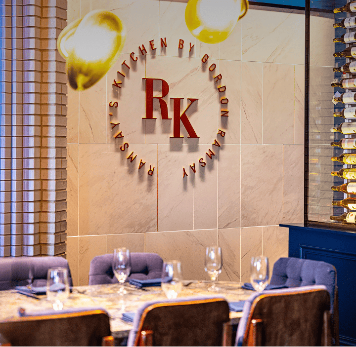 Elegant dining area with laid table, wine glasses, wall sign “Ramsay’s Kitchen by Gordon Ramsay,” and wine display.
