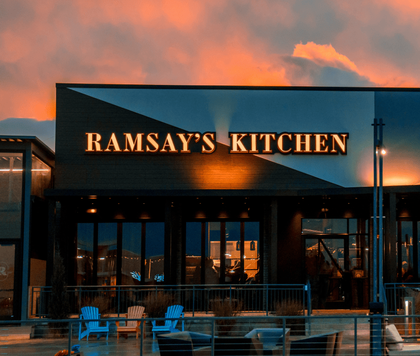 Restaurant exterior at sunset with sign reading “Ramsay’s Kitchen” and outdoor seating in the foreground.