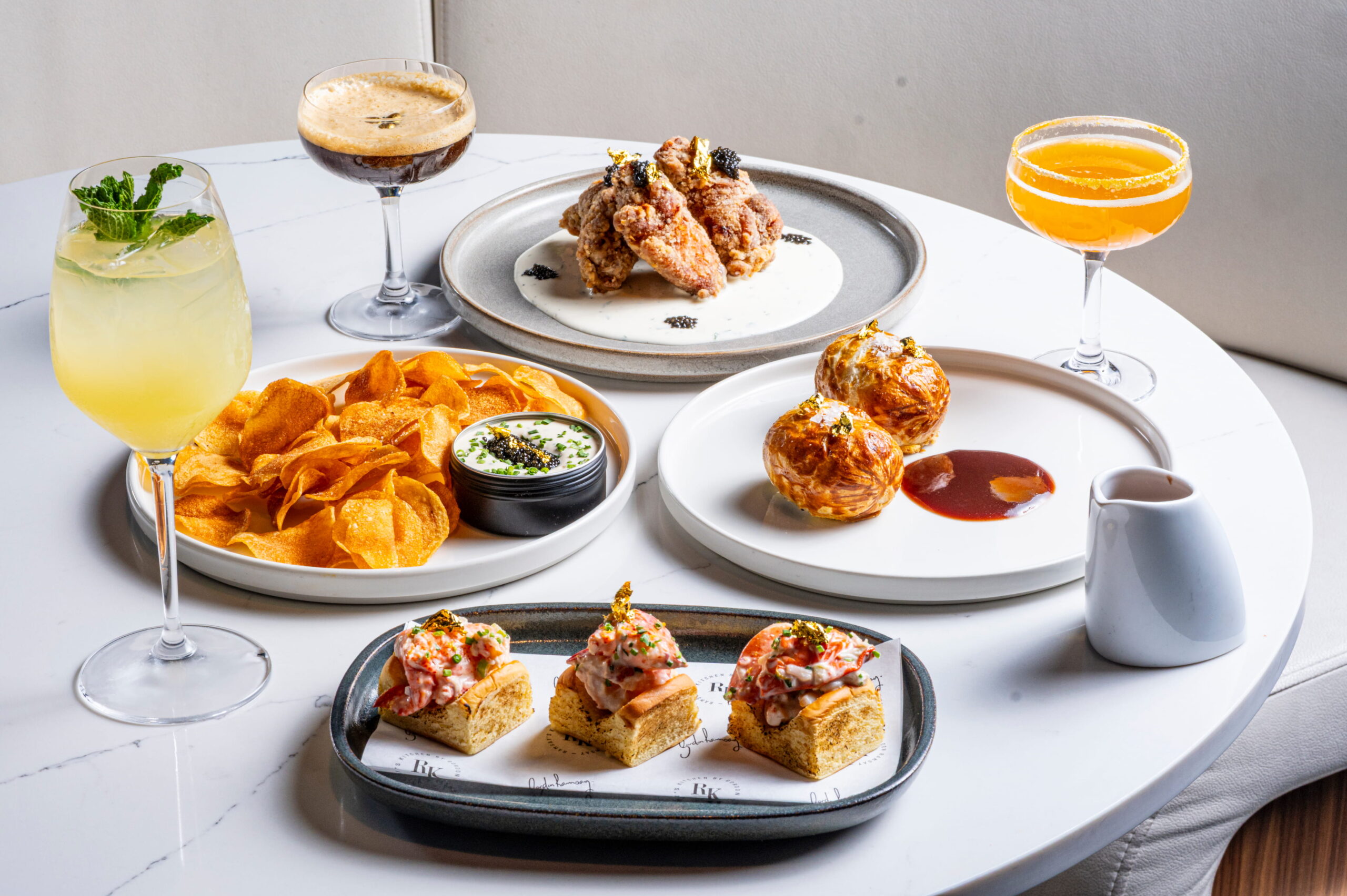 A table with cocktails, crisps with dip, fried chicken, pastries with sauce, and topped bread appetisers.
