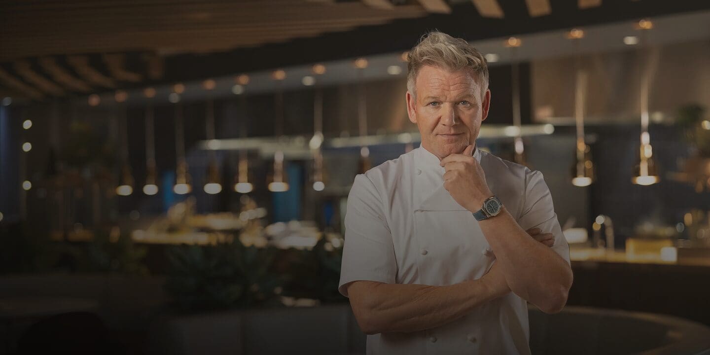 A chef in a white uniform stands in a modern restaurant, looking seriously at the camera.