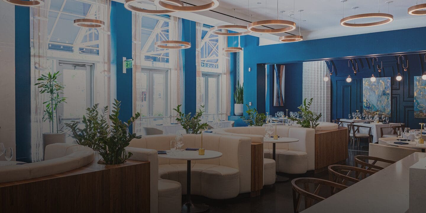 Modern restaurant with blue walls, white cushioned seating, round lights, and plants on tables.