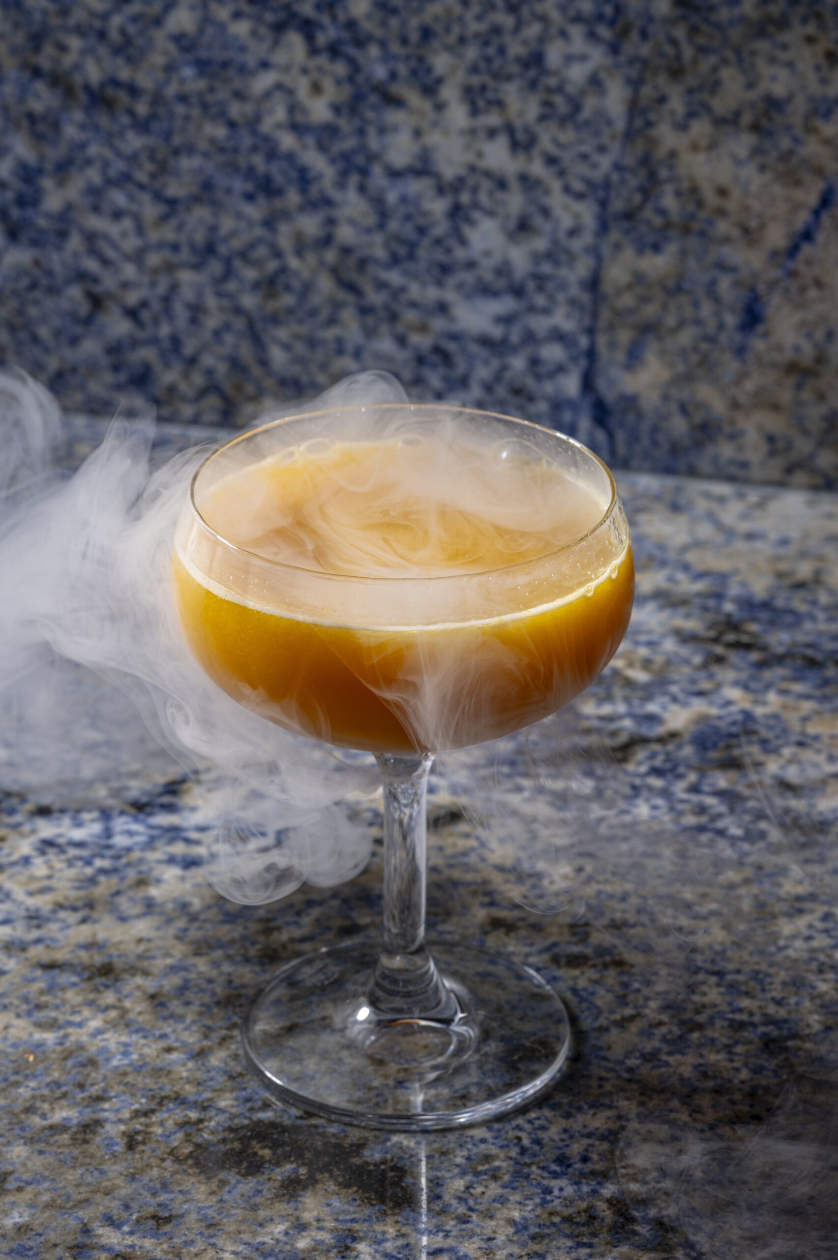 A stemmed glass filled with an orange cocktail surrounded by dry ice vapour.