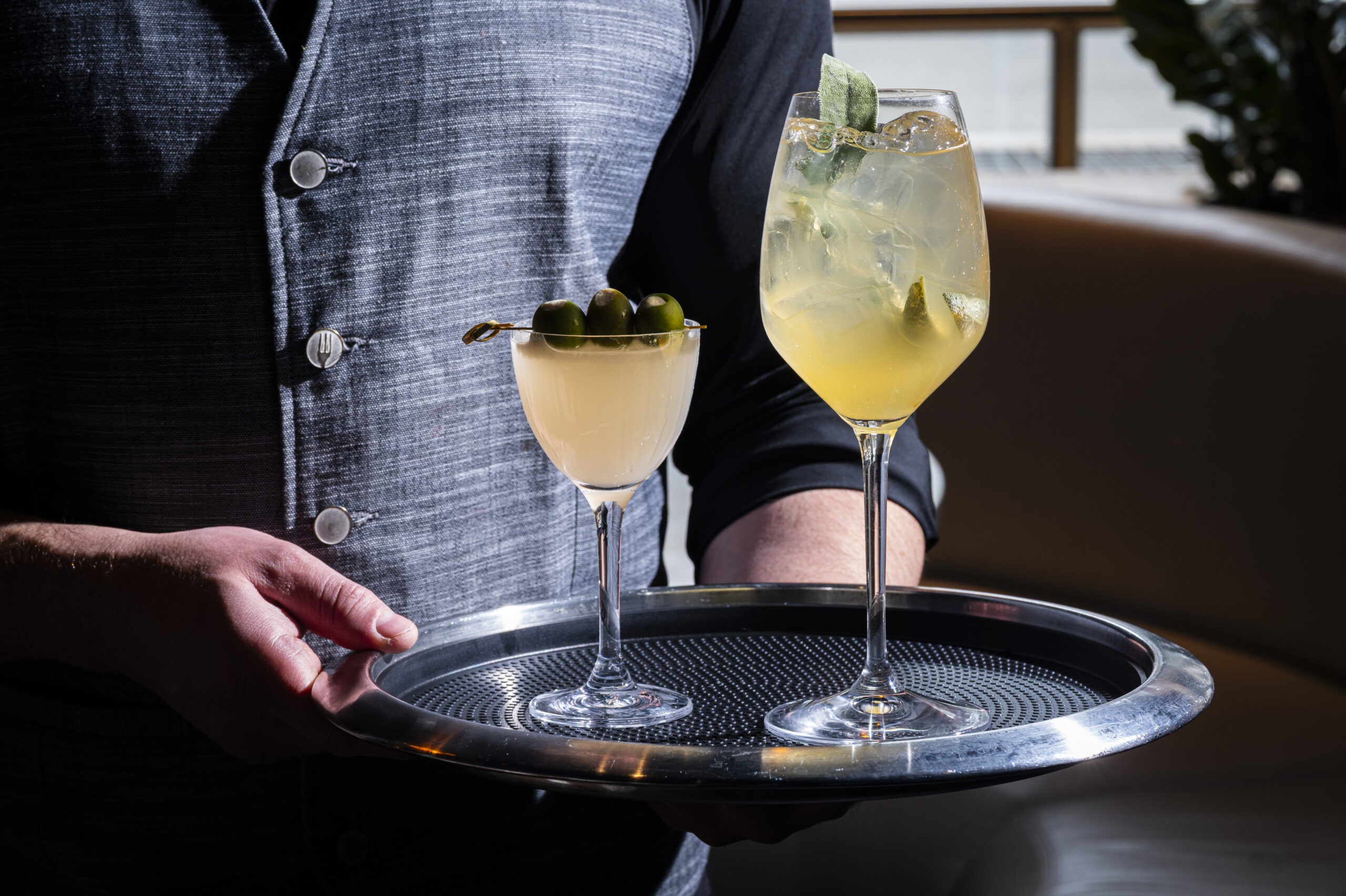 A person in a waistcoat holding a silver tray with a martini garnished with olives and a cocktail with a herb garnish.