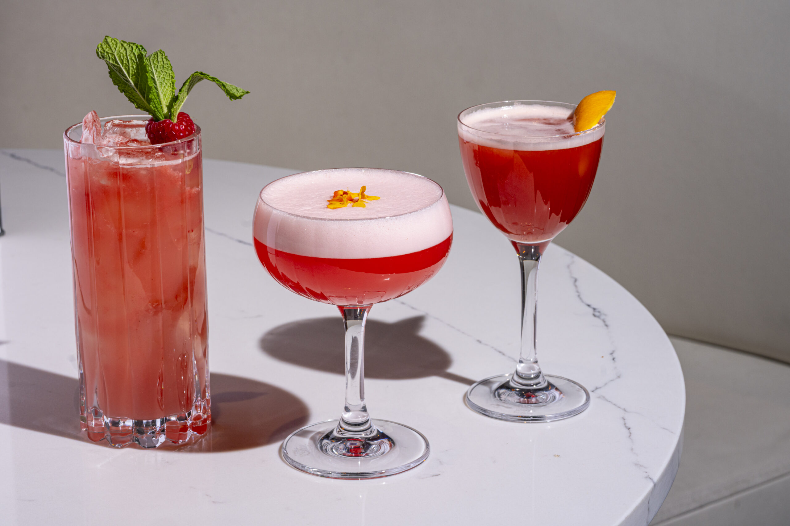 Three colorful red cocktails served in different glassware on a white marble table.