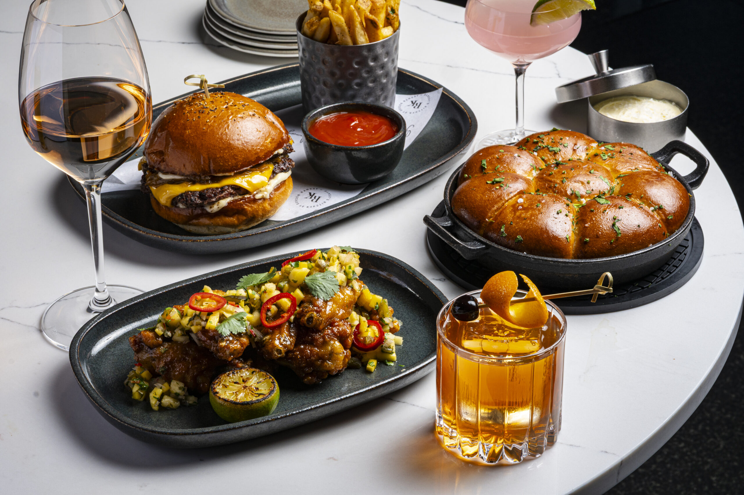 A spread of restaurant food including a cheeseburger, chicken wings, pull-apart bread, chips, and three cocktails.