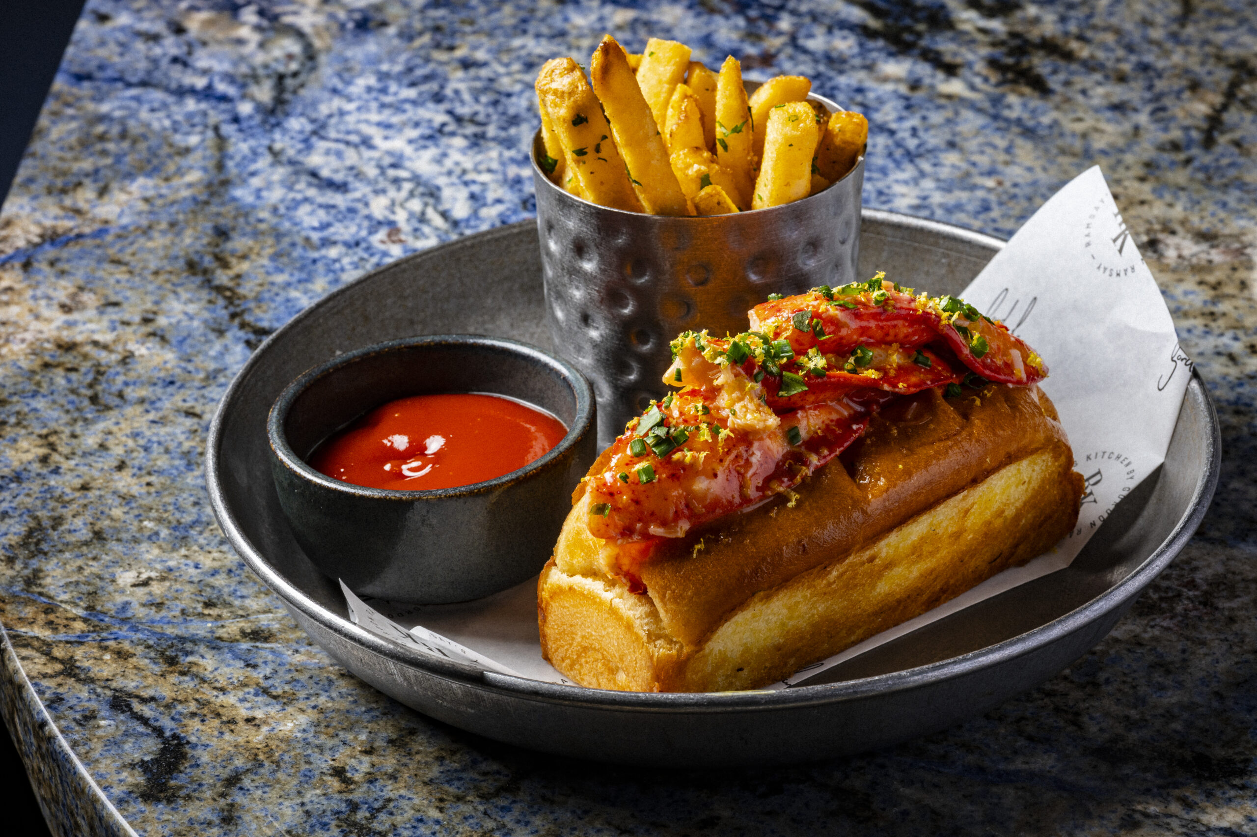 A lobster roll in a bap served with a side of chips and a small bowl of tomato sauce.