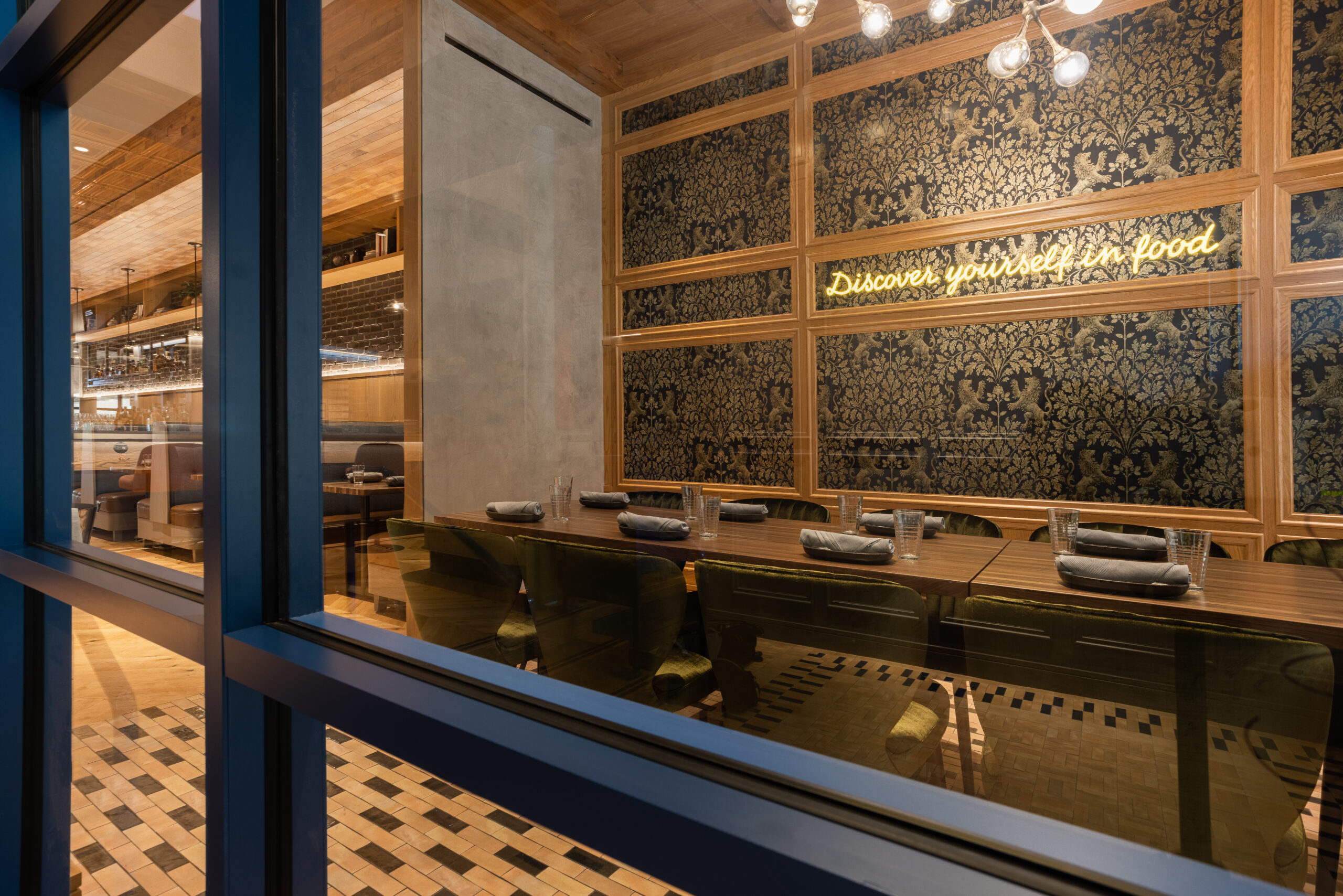 Dining area seen through a window with a neon sign reading “Discover yourself in food” on a patterned wall.