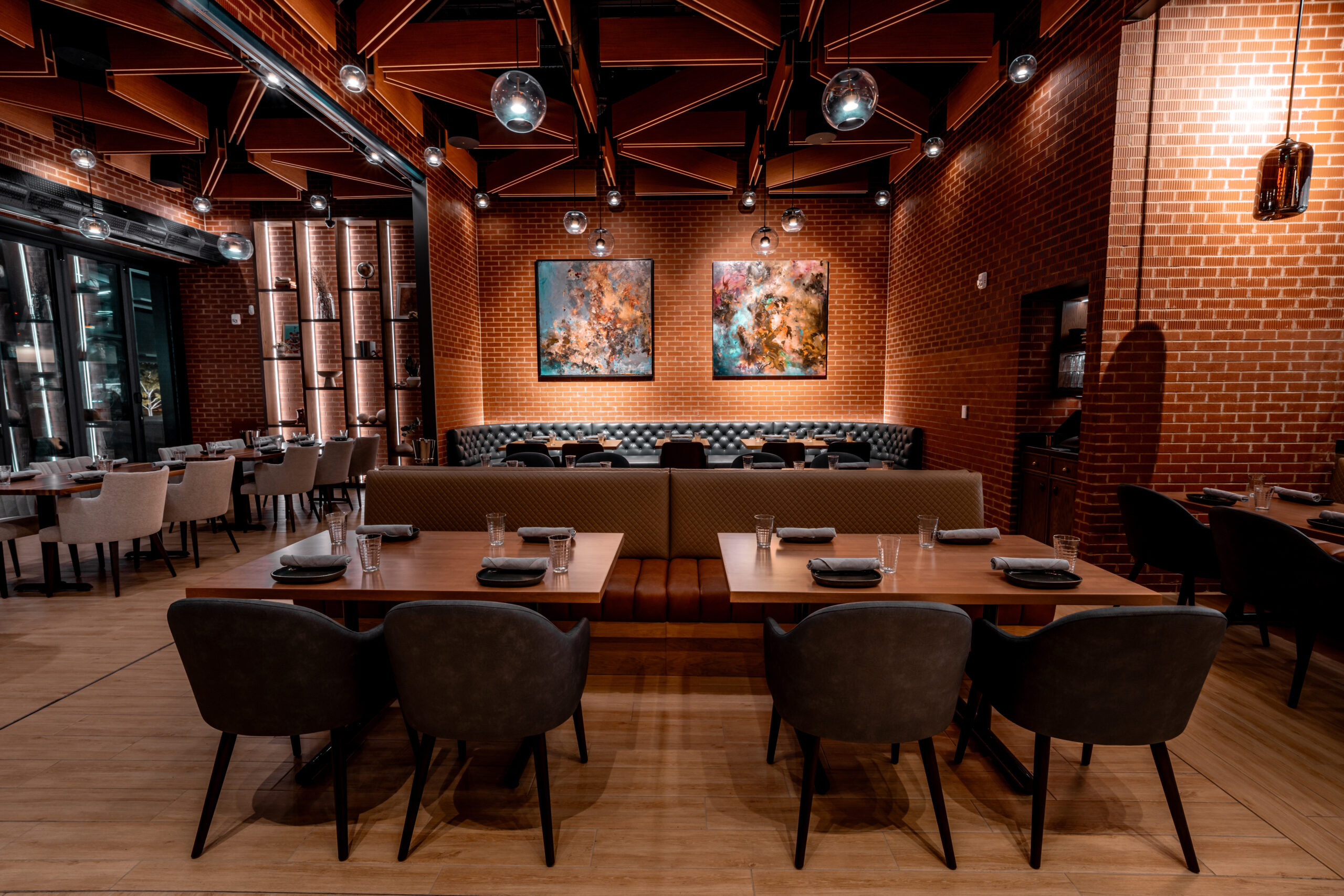 Dining room with two booths facing each other, laid tables, and two abstract paintings on a brick wall.