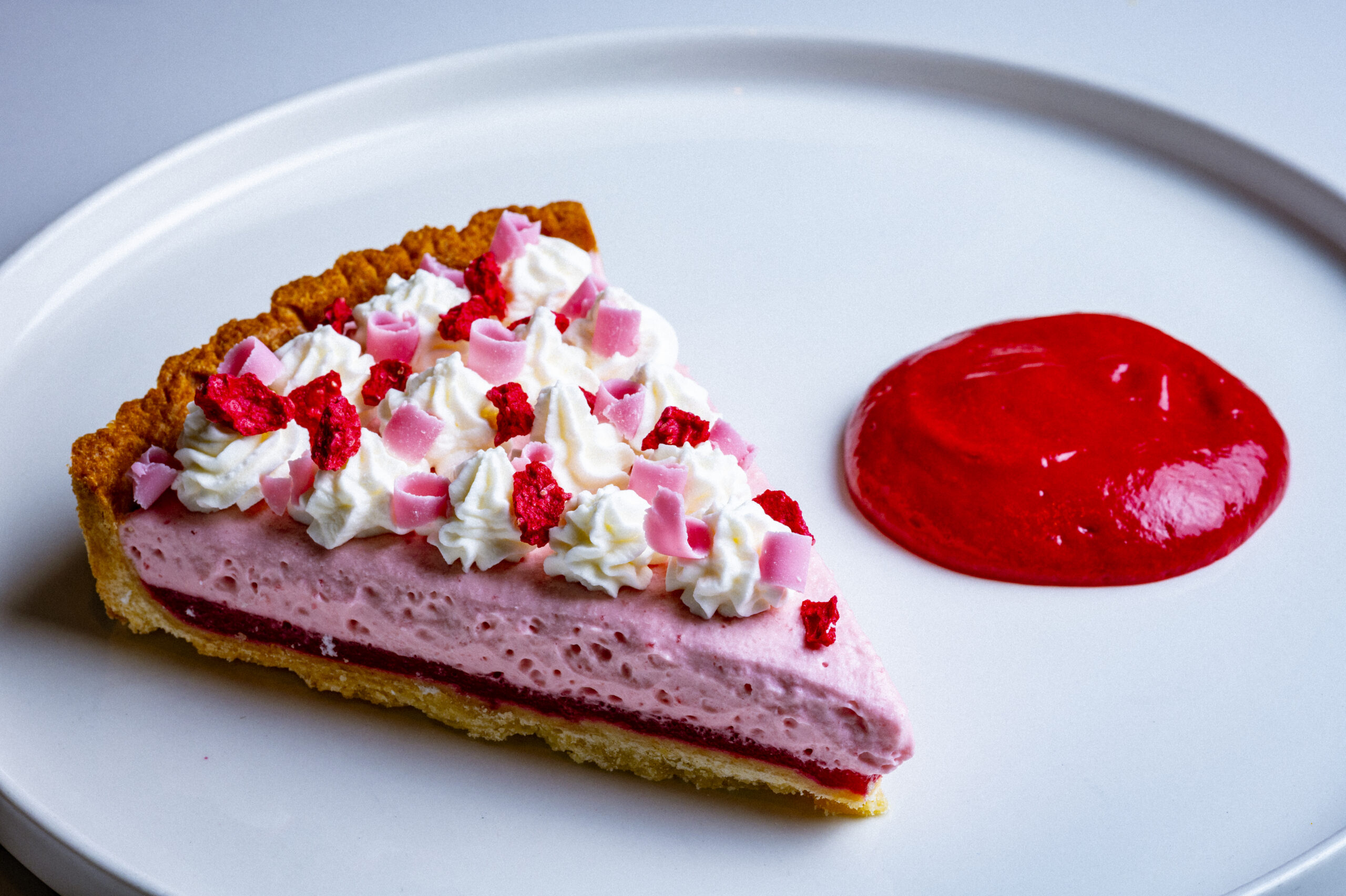 A slice of pink berry tart topped with whipped cream and chocolate curls, served with a dollop of fruit coulis.