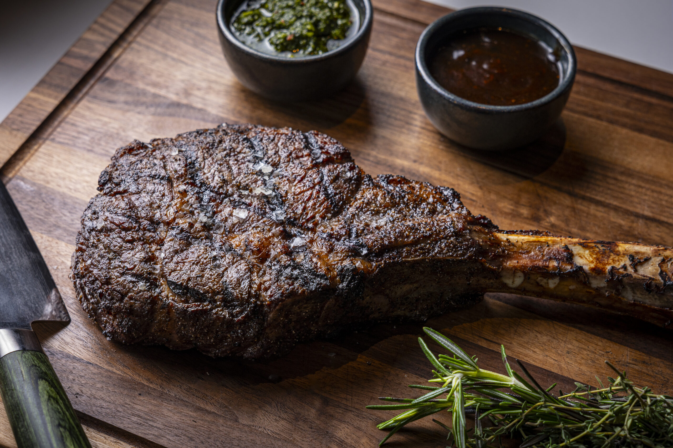 A grilled tomahawk steak on a wooden chopping board with two small bowls of sauce and a sprig of rosemary.