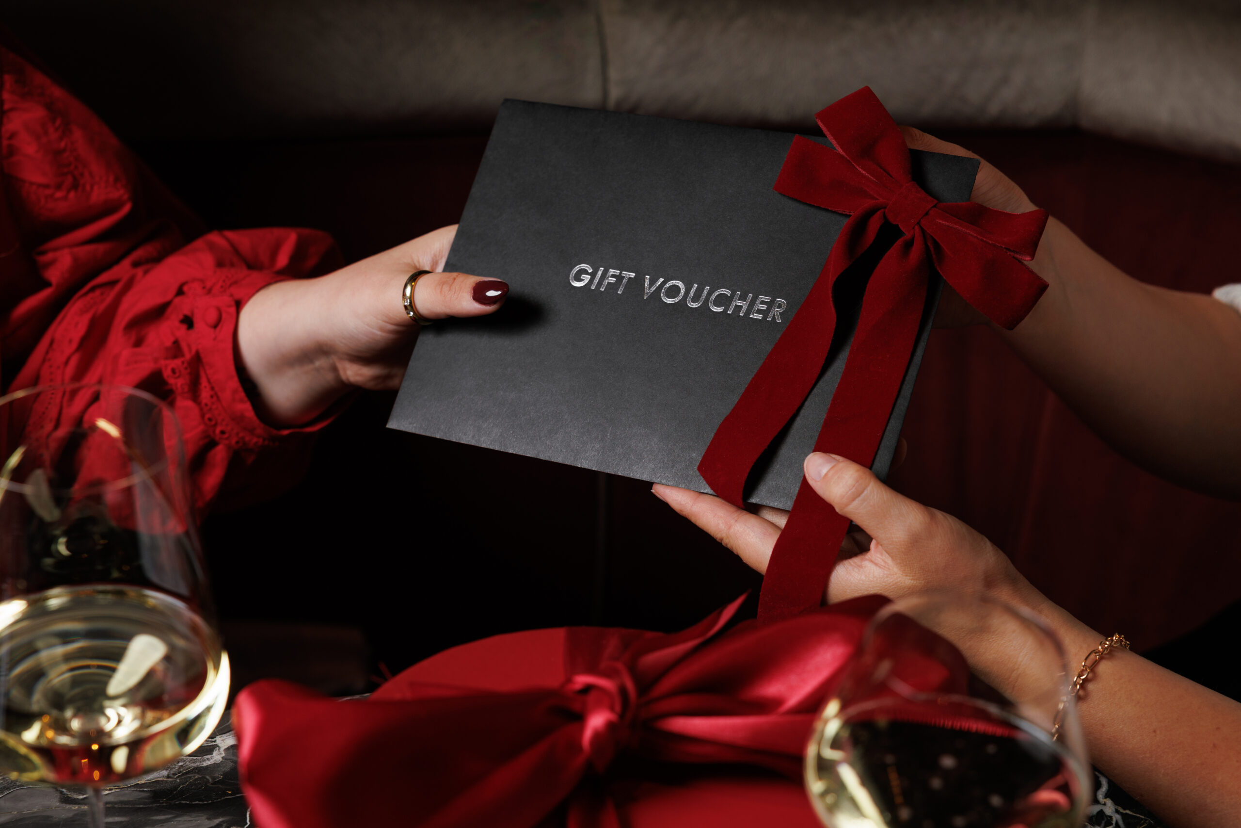 Two hands exchange a black gift voucher envelope tied with a red velvet ribbon.