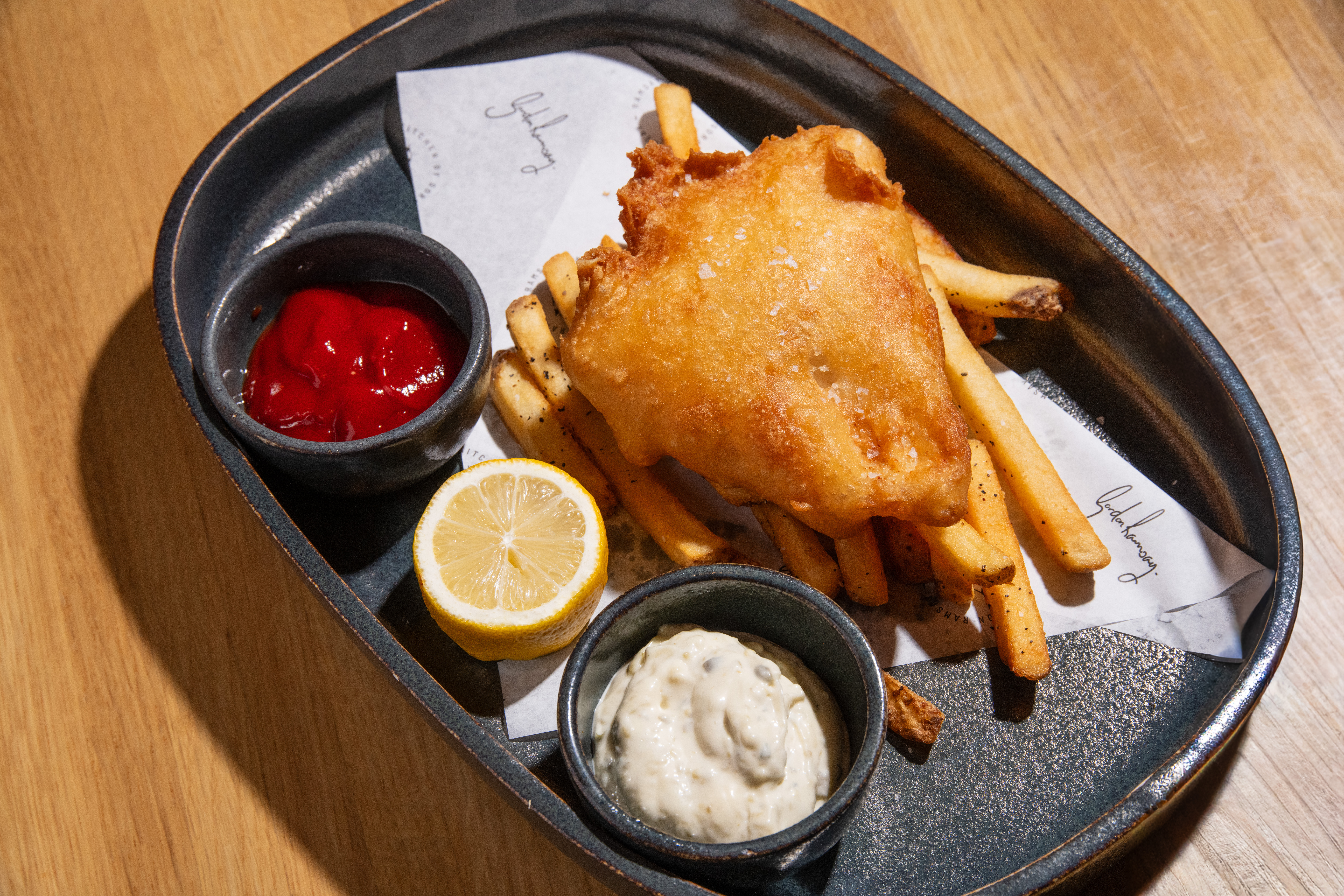 Fish and chips with chips, tartare sauce, tomato ketchup, and a lemon wedge on a black plate on a wooden table.