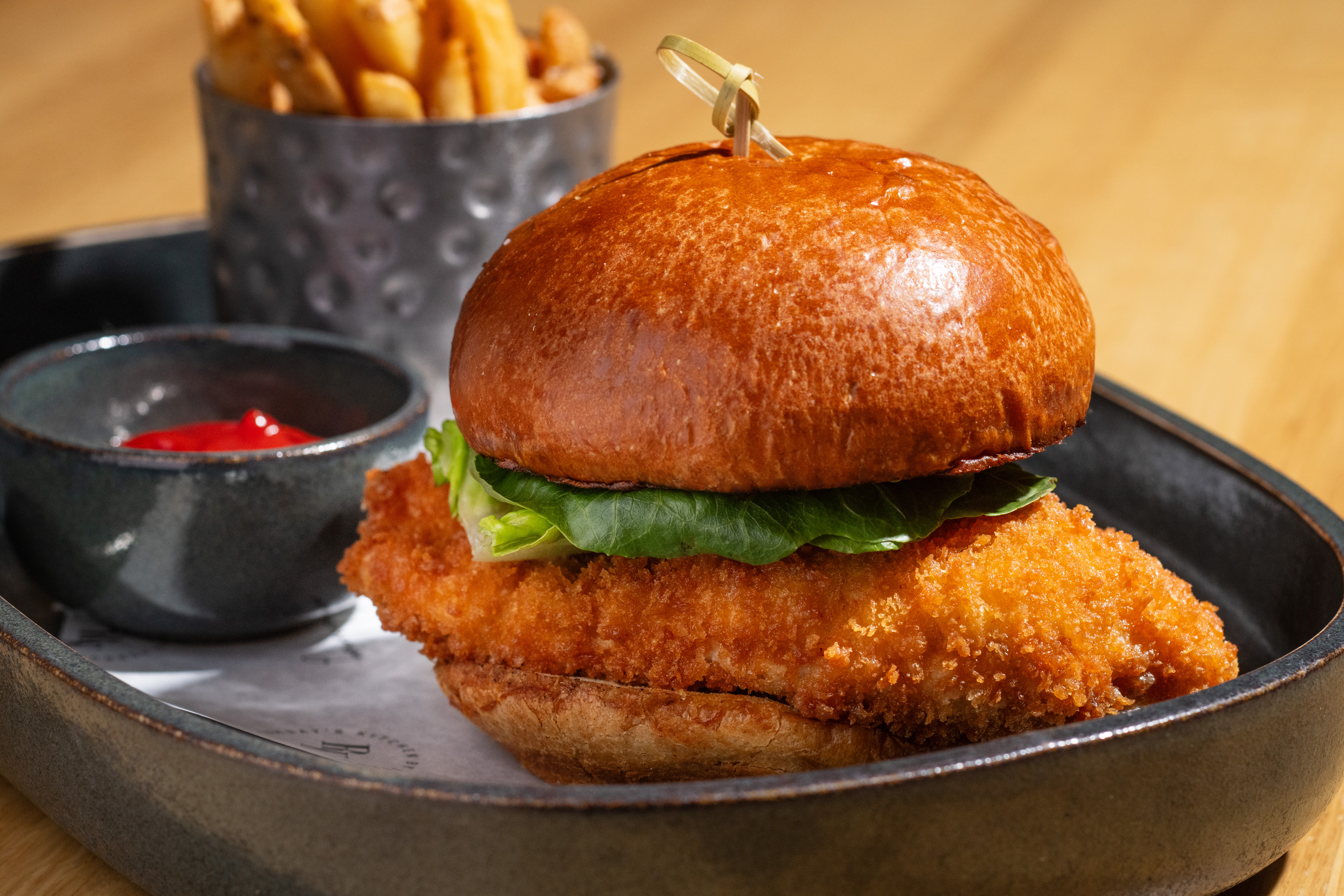 Crispy chicken sandwich with lettuce in a bap, served with chips and tomato sauce on a tray.