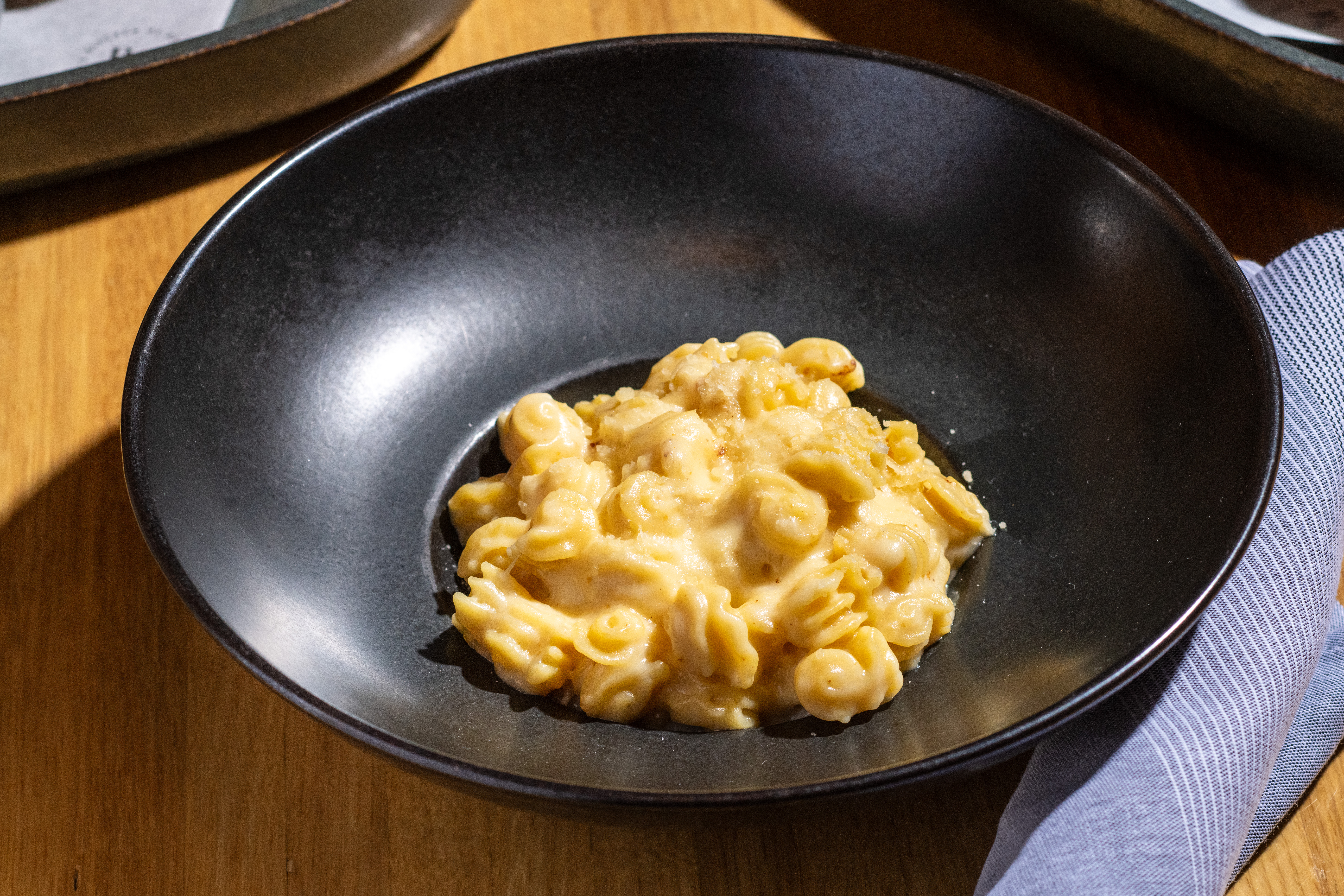 Creamy macaroni cheese served in a black bowl on a wooden table with a blue cloth serviette.