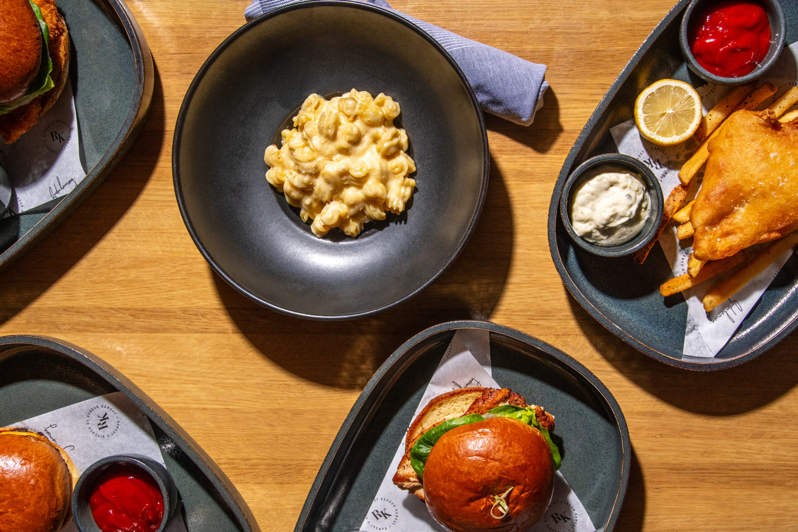 Overhead view of bowls of macaroni cheese, fish and chips, and chicken sandwiches on a wooden table.