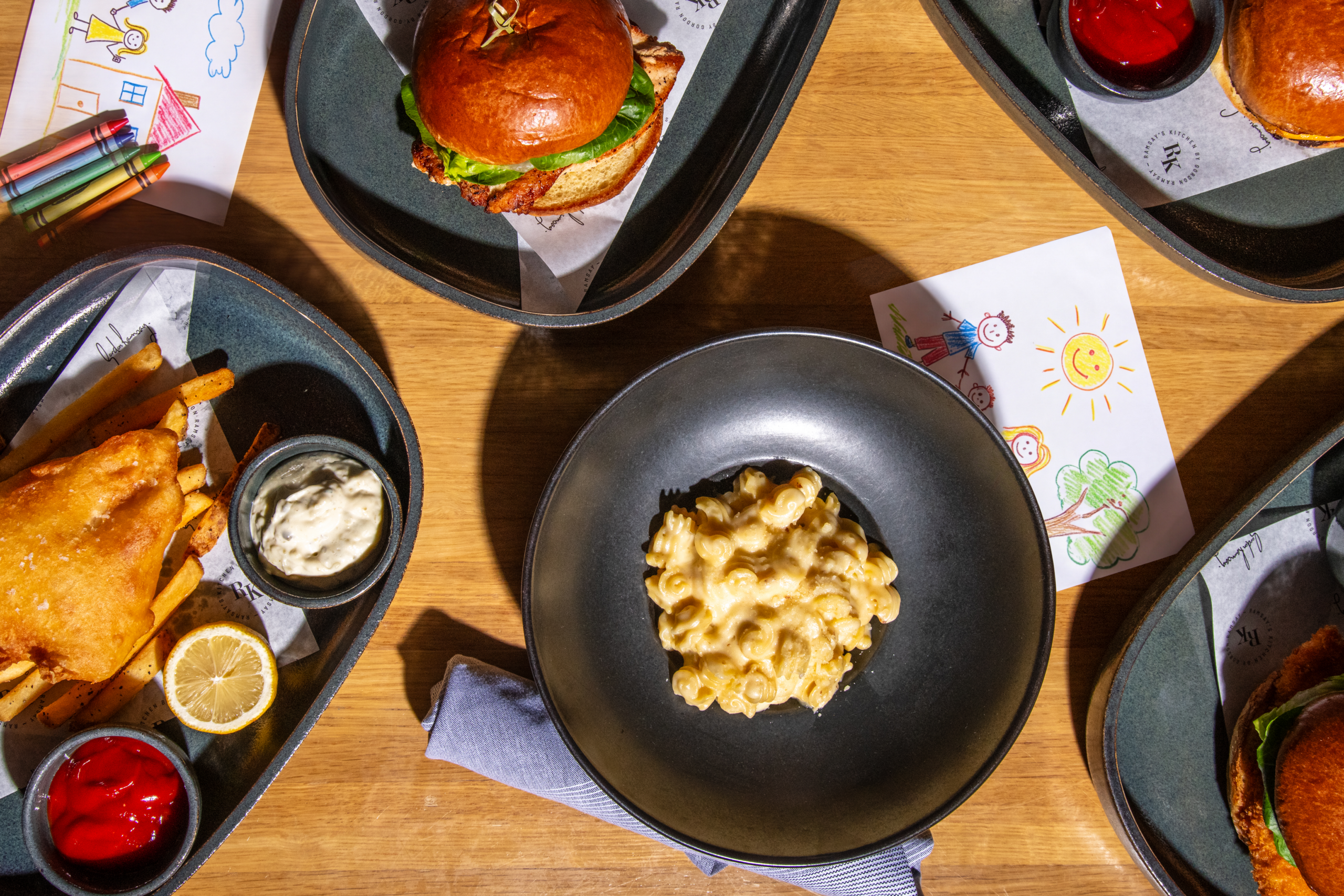 A flat lay of restaurant meals including a burger, fish and chips, and macaroni cheese with children's drawings.