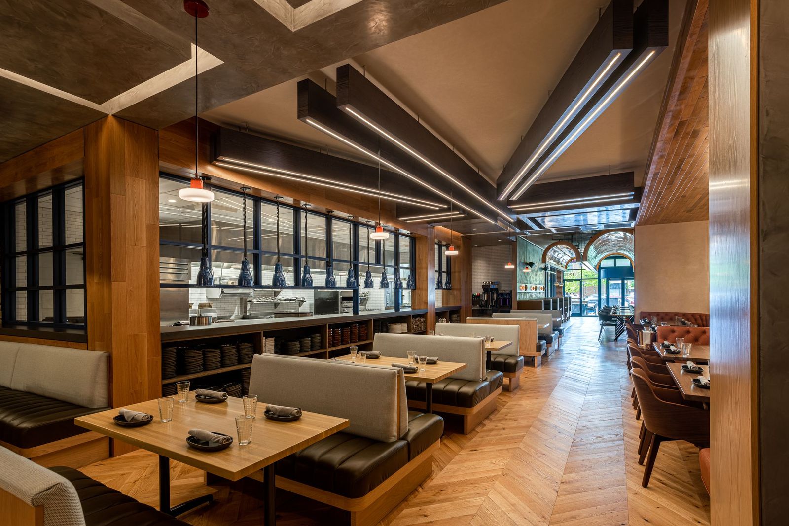 Interior of a restaurant with wooden booths, tables set with serviettes and glasses, and an open kitchen.