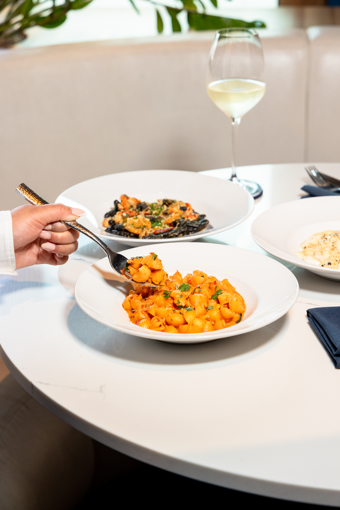 A person lifts a forkful of orange pasta from a bowl, with other dishes and a glass of white wine on the table.