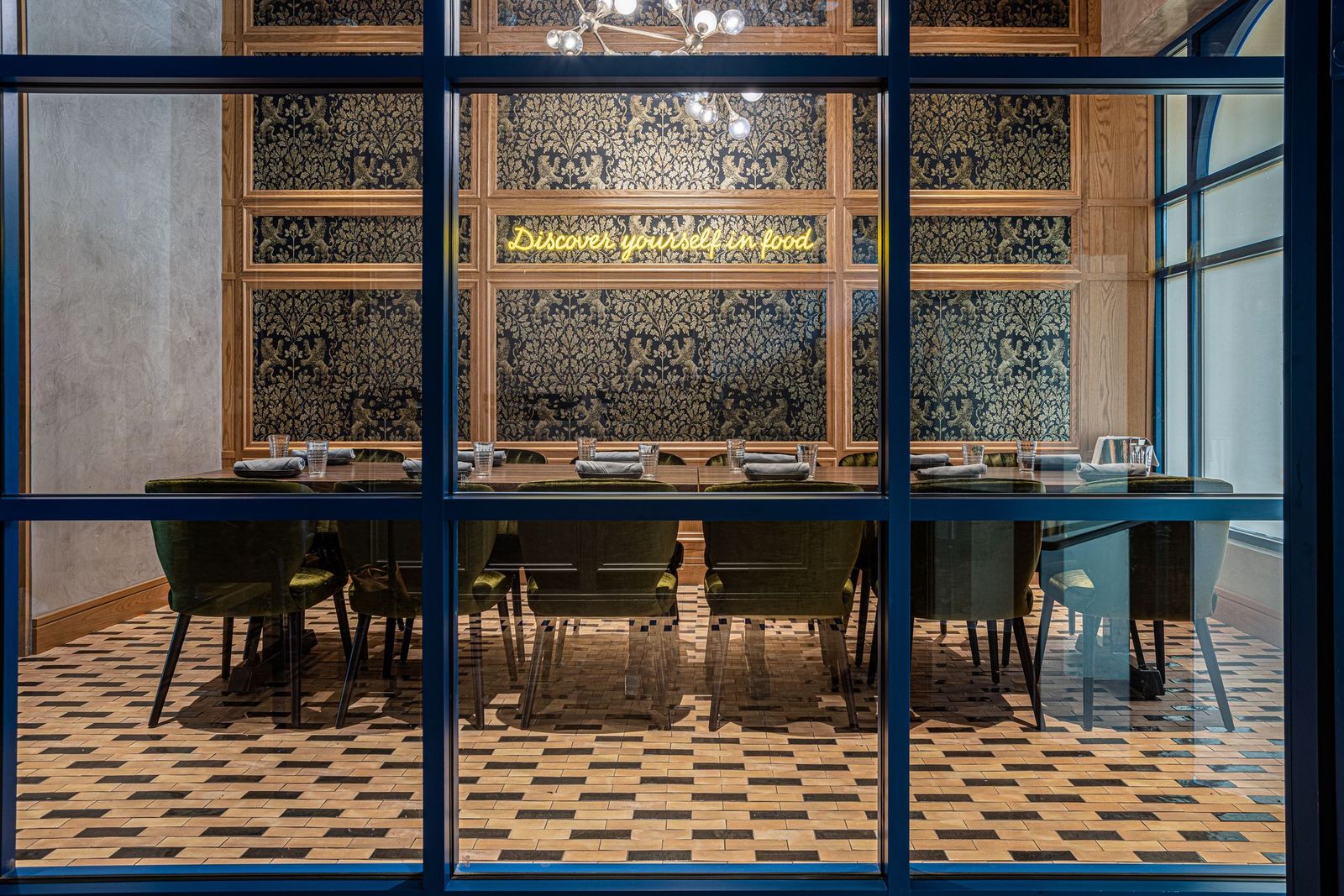 A long dining table with green chairs set in front of a wall with a neon sign reading “Discover yourself in food.”