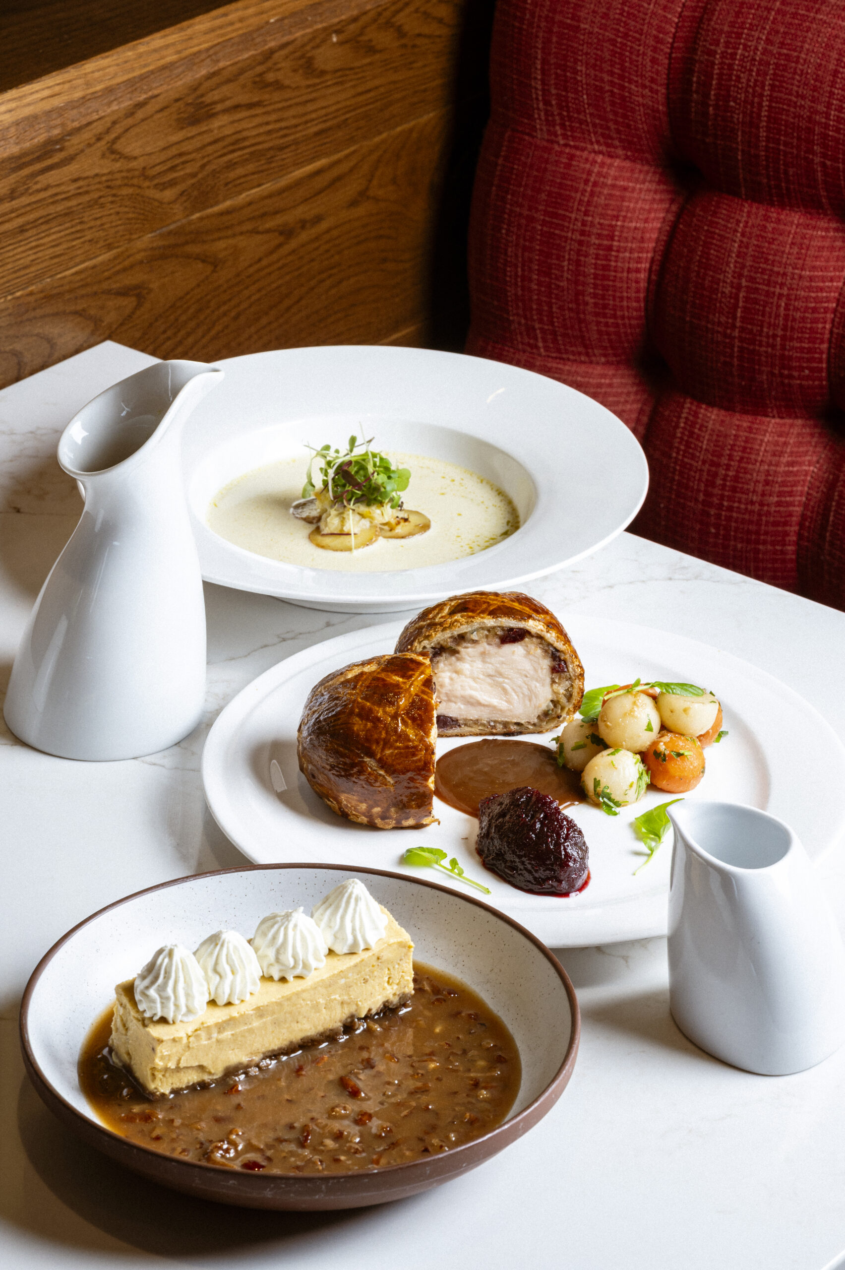 A bowl of cream soup, a plate of beef Wellington with vegetables, and a slice of pudding with whipped cream.